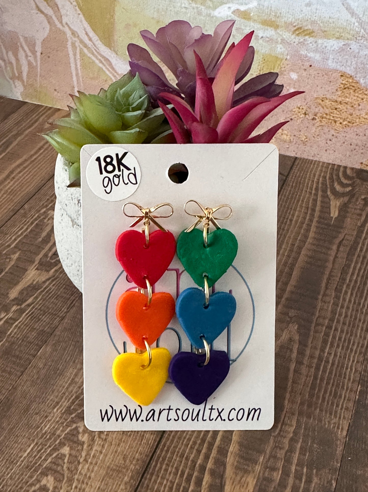 Colorful heart-shaped earrings on a branded card with '18K Gold' and website text, set against a wooden surface.