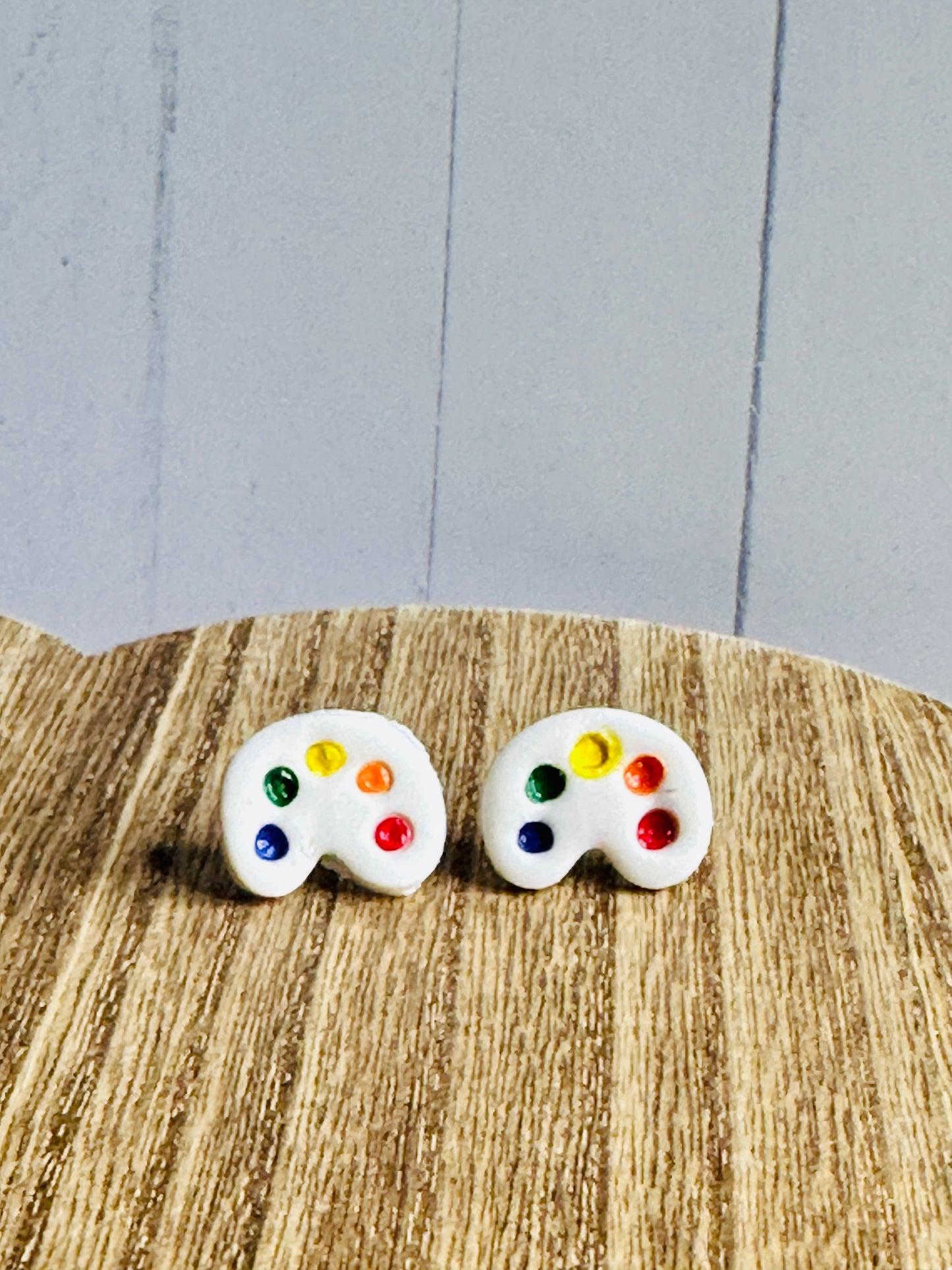 A pair of art palette stud earrings on a wooden table. The palette is white and has dots of blue, green, yellow, orange, and red paint.