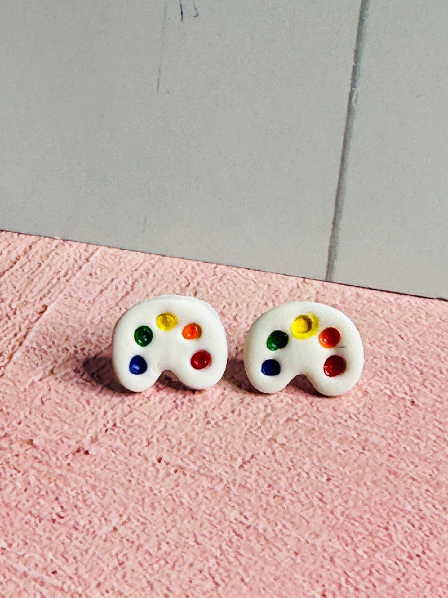 A pair of art palette stud earrings on a pink table in front of a white wall. The palette is white and has dots of blue, green, yellow, orange, and red paint.