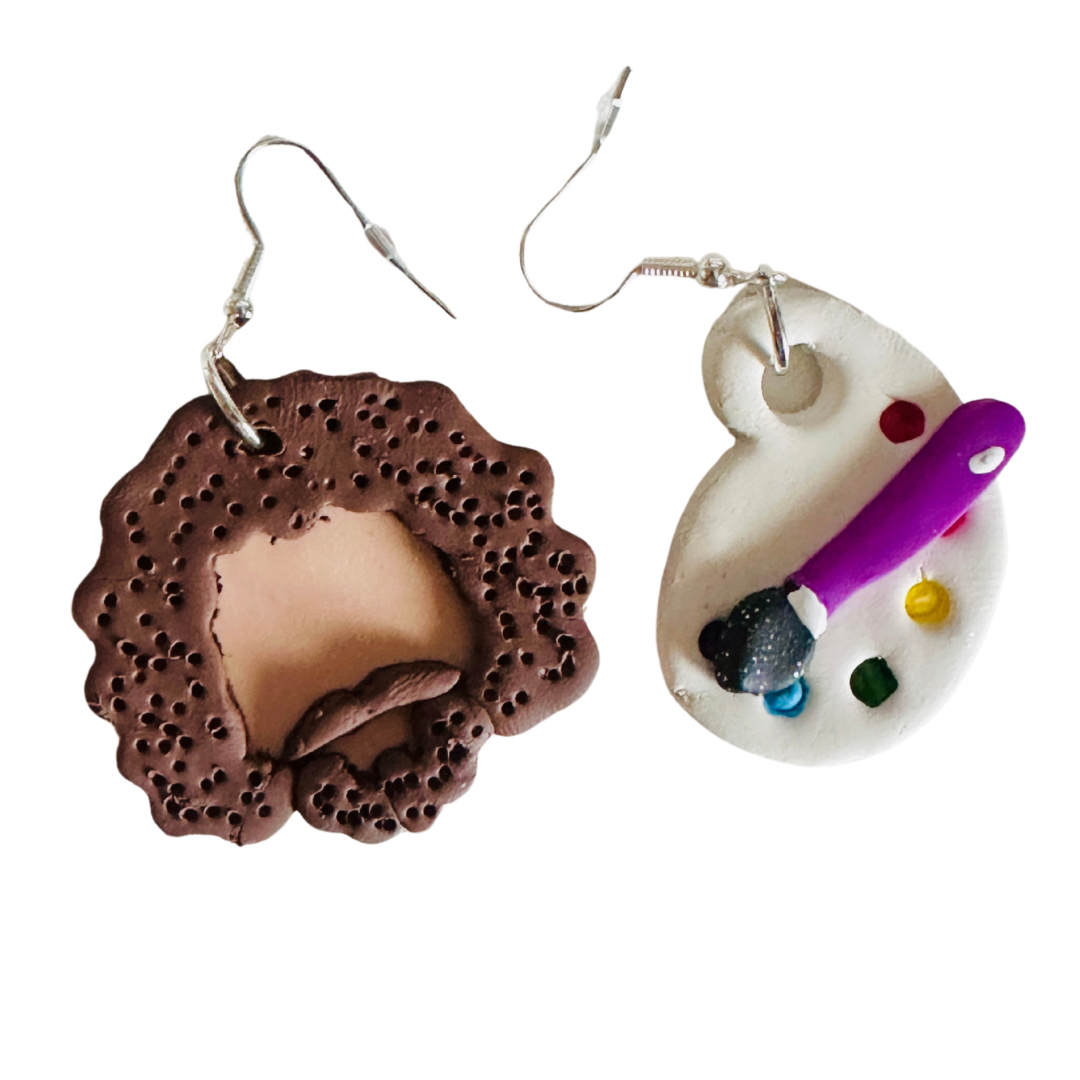 Handmade Bob Ross Style Polymer Clay Earrings. The earring on the left is a 1.5 x 1.5 inch Bob Ross Head shaped with beige skin, brown hair, beard, and mustache. The earring on the right is a 1 x 1.4 inch paint palette with a paintbrush on top. The paint palette has blue, green, red, and yellow paint dots.
