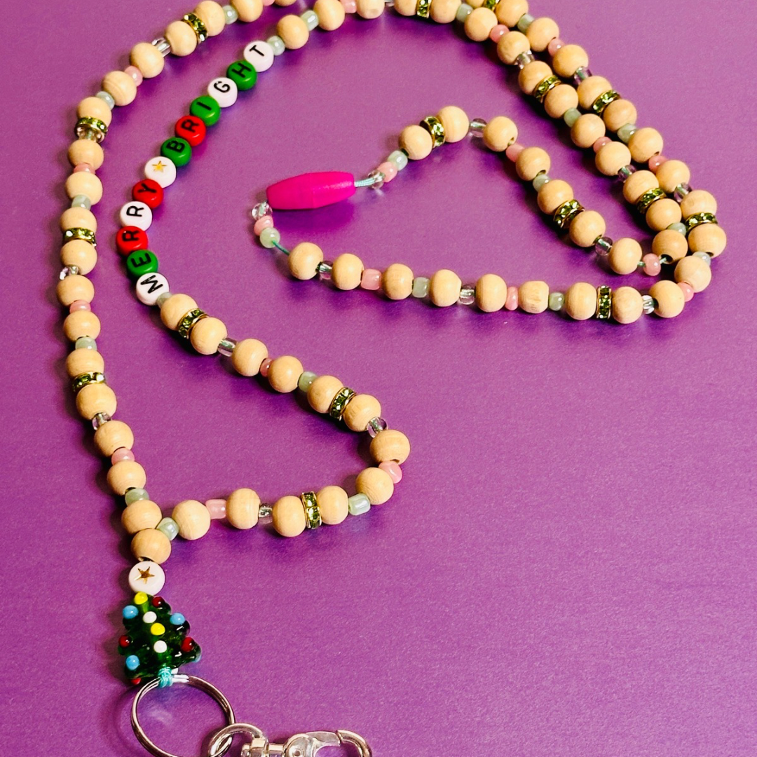 The Christmas Tree Lanyard viewed from an angle with the Christmas Tree charm on the bottom left, the beaded words "MERRY AND BRIGHT" on the upper left, and the pink breakaway clasp in the middle. The lanyard is laying in a circular shape.