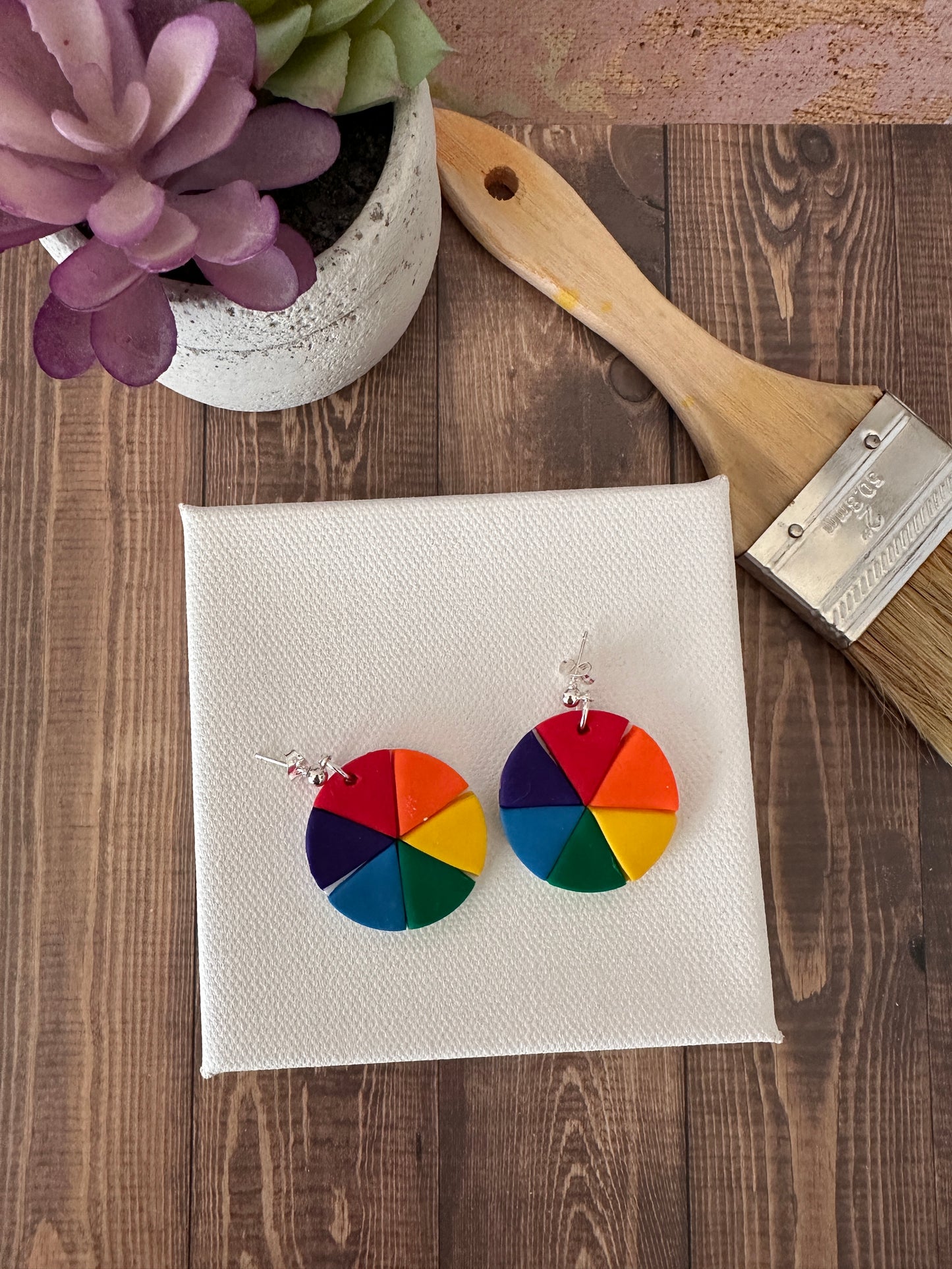 Top-down view of color wheel polymer clay earrings resting on a white artist canvas next to a paintbrush.