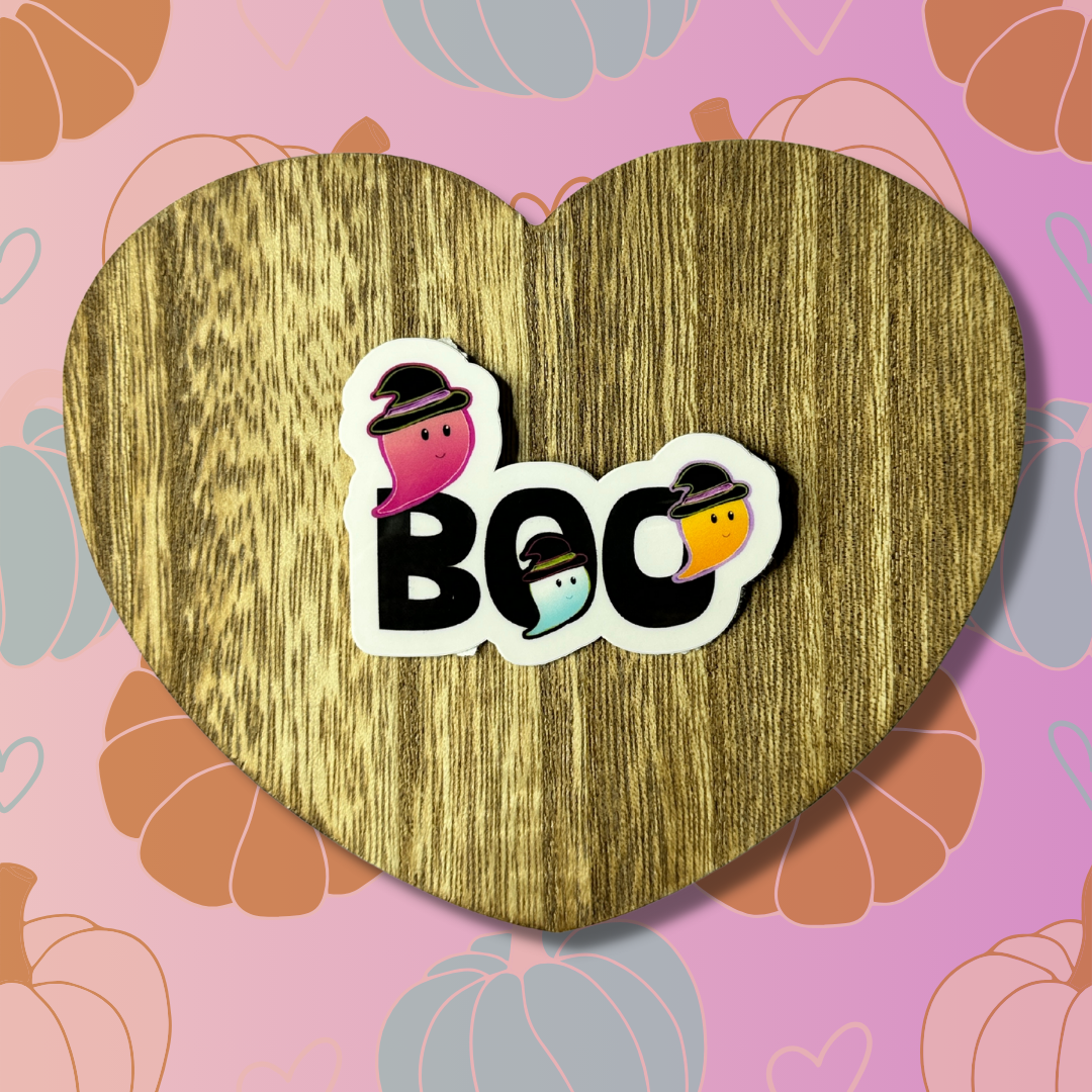 Cute ghosts Halloween vinyl sticker displayed on heart-shaped wooden table with pink and orange background showing three colorful ghosts saying BOO