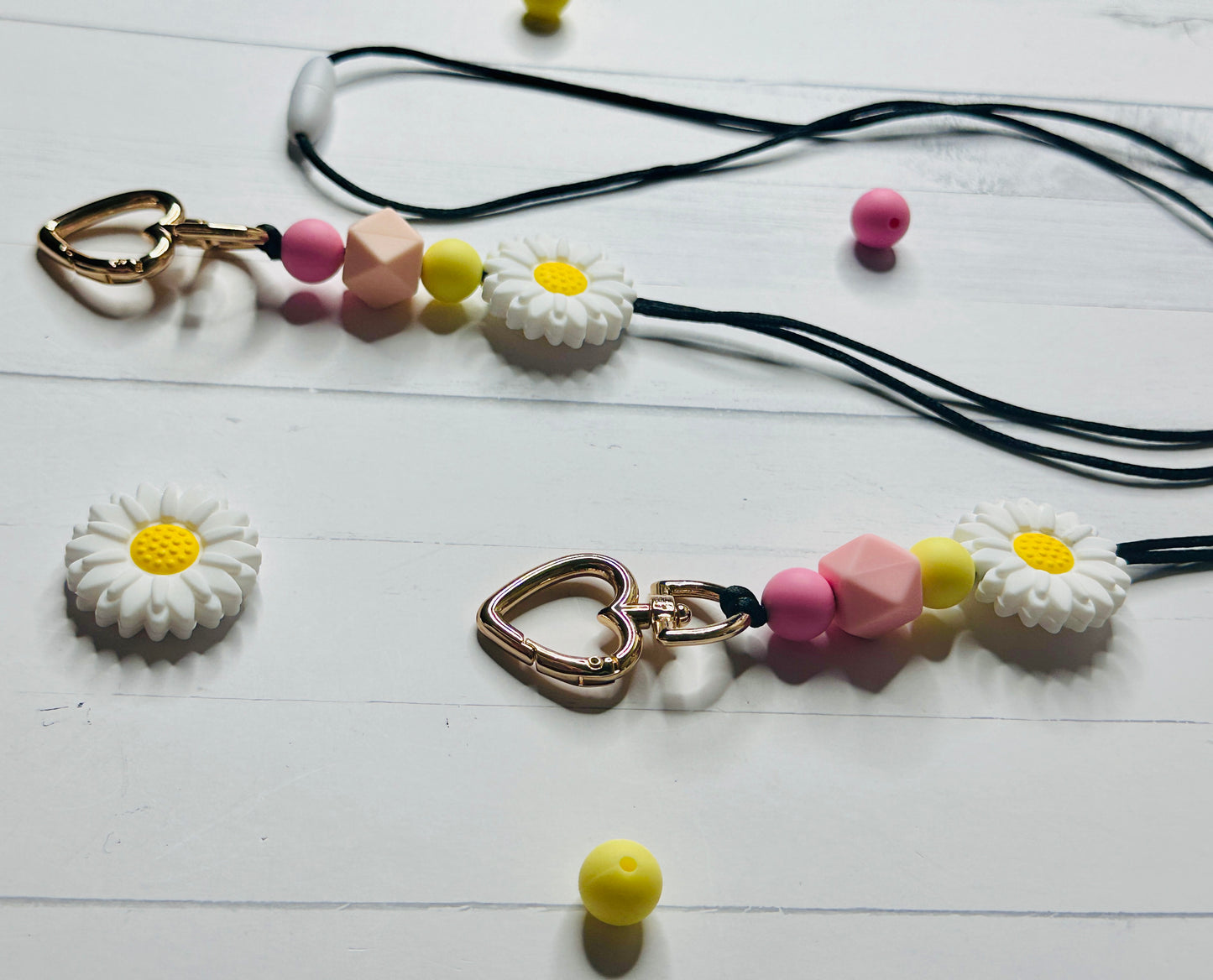 A close up and angled view of the Flower Lanyard. The heart shaped metal clasp, silicon beads, and daisy flower are displayed on a white wooden table and are casting shadows onto its surface.