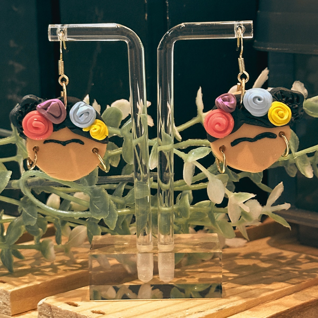 A pair of Frida Kahlo earrings hanging from a clear earring stand. Green plants are behind the earrings and casting a shadow on the table.