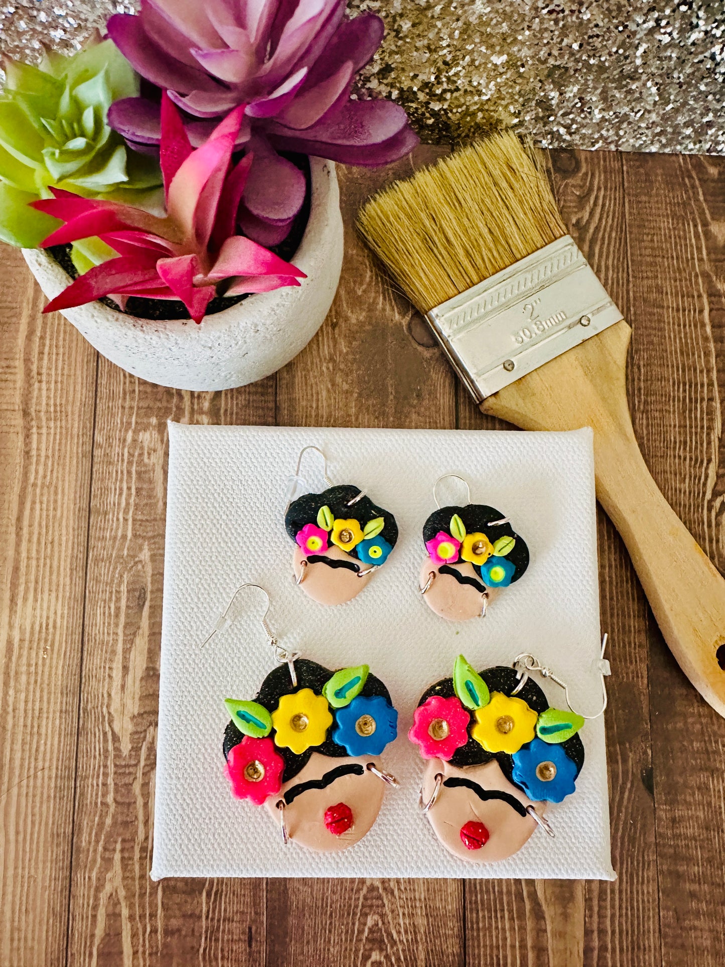 Large and small variants of Frida Kahlo-themed earrings on a white canvas with a brush and plant in the background.