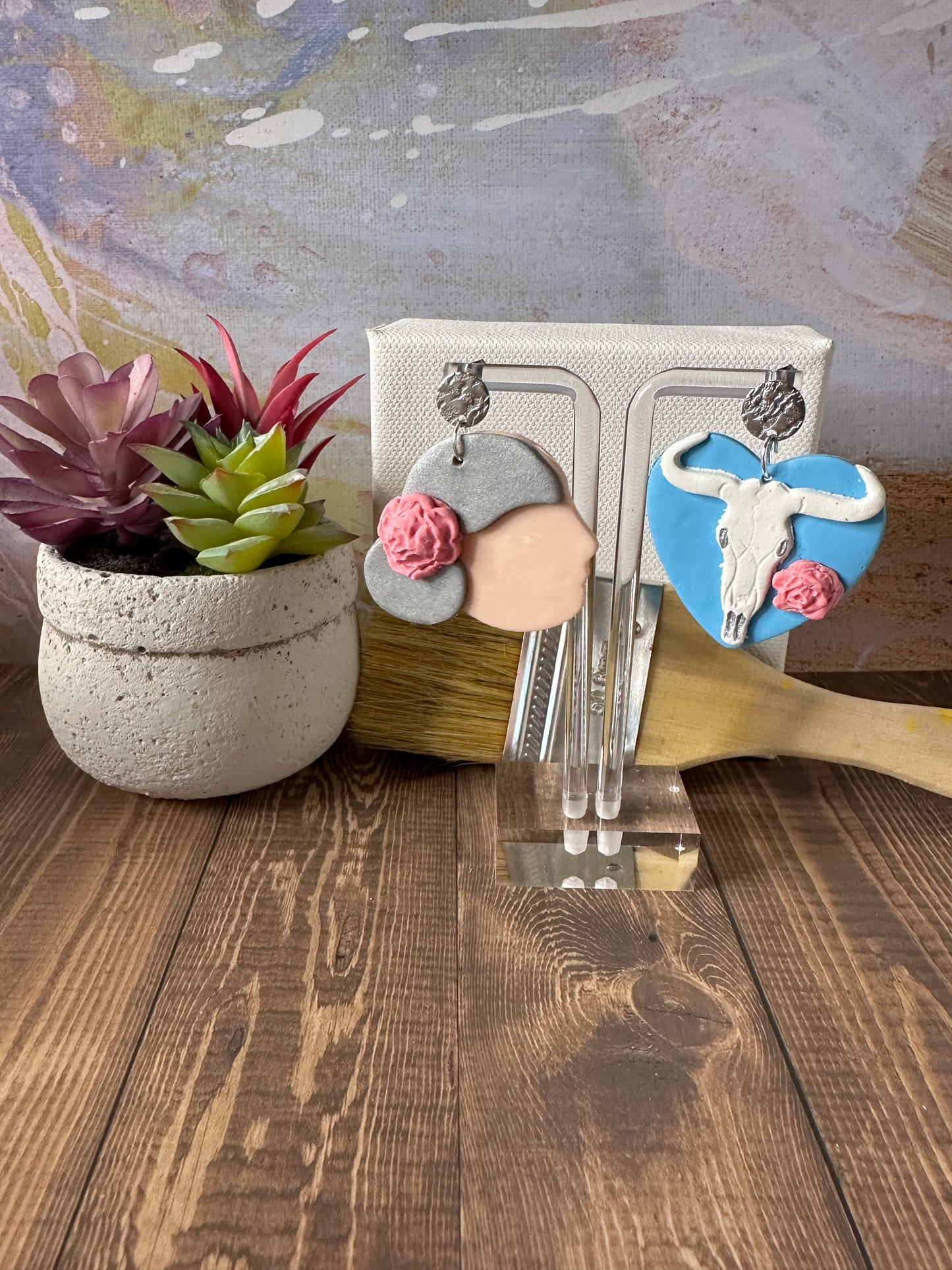 Georgia O'Keeffe polymer clay earrings hanging from an earring stand with a flower pot, mini canvas, and paintbrush styled in the background.
