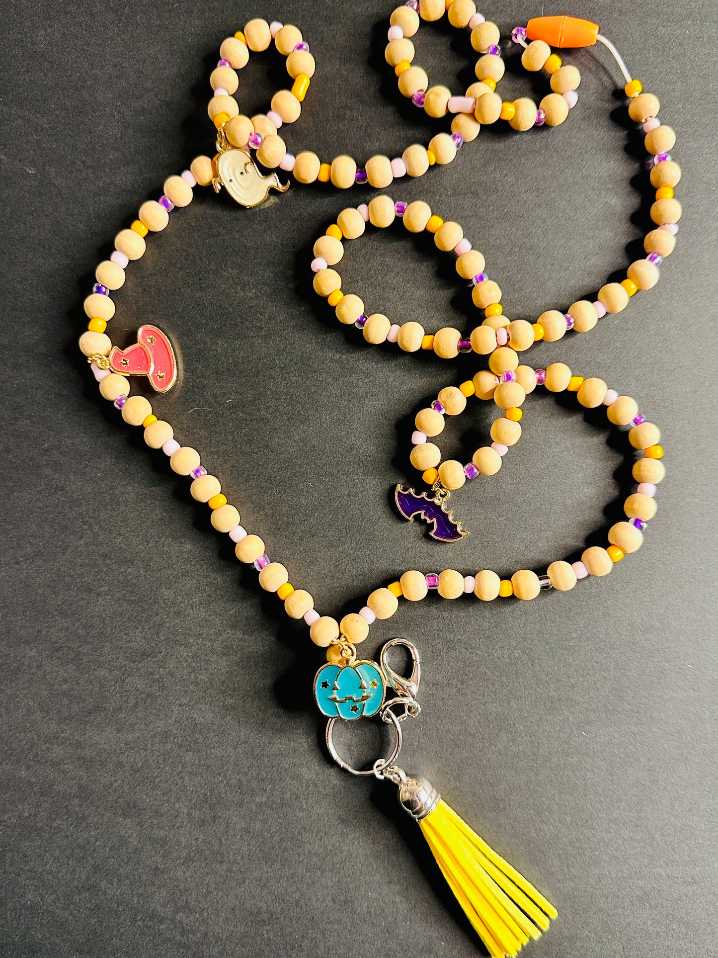 The Halloween Lanyard on a grey background. The full lanyard can be seen with its wooden beads, metal charms shaped like pumpkins, bats, witch hats, and ghosts, and colorful tassel.