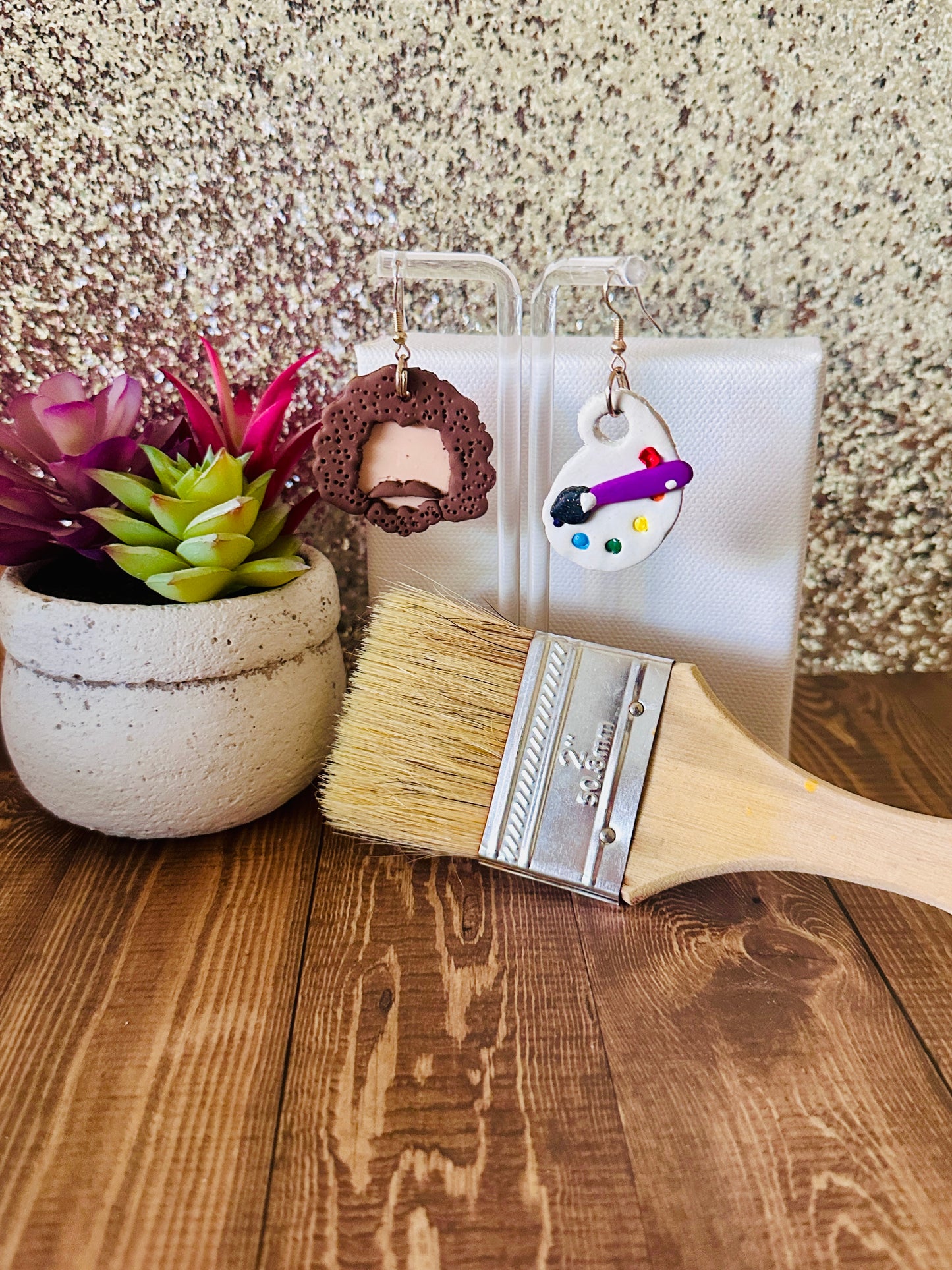 Large Bob Ross earring set displayed by a paint canvas with a paintbrush and a plant on a wooden surface.