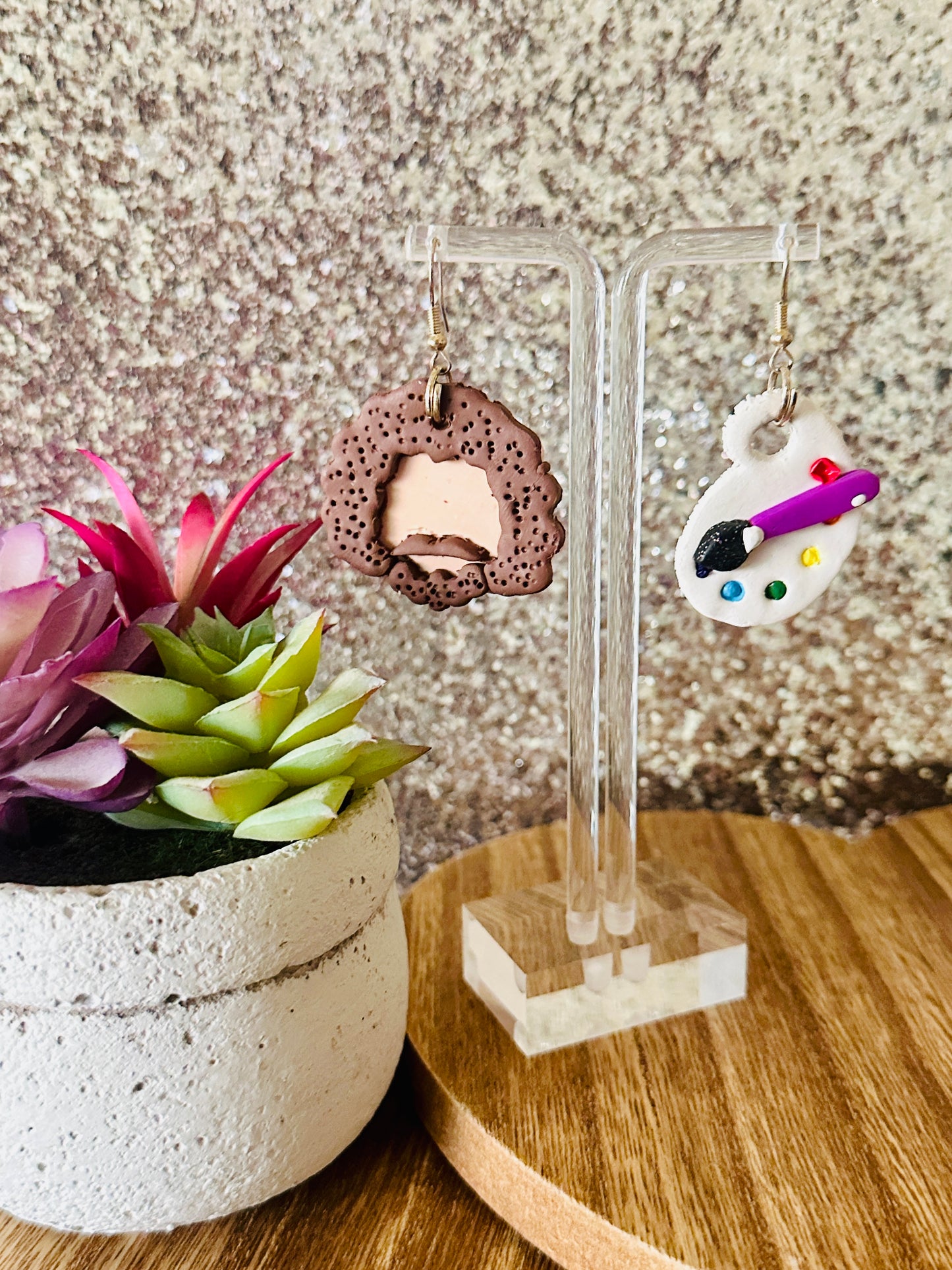 The large variant of the Bob Ross polymer clay earrings on a clear stand with a potted plant and glittery background.