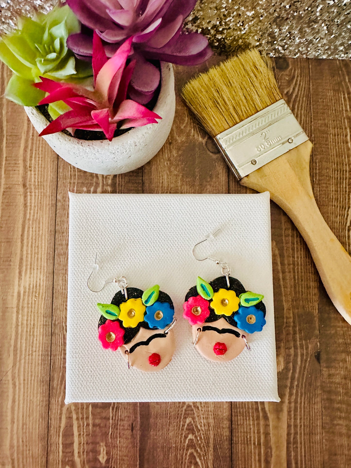 The large variant of Frida Kahlo-themed earrings with red lips on a white artist canvas with a paintbrush and flowers in the background and sitting on a wooden table.