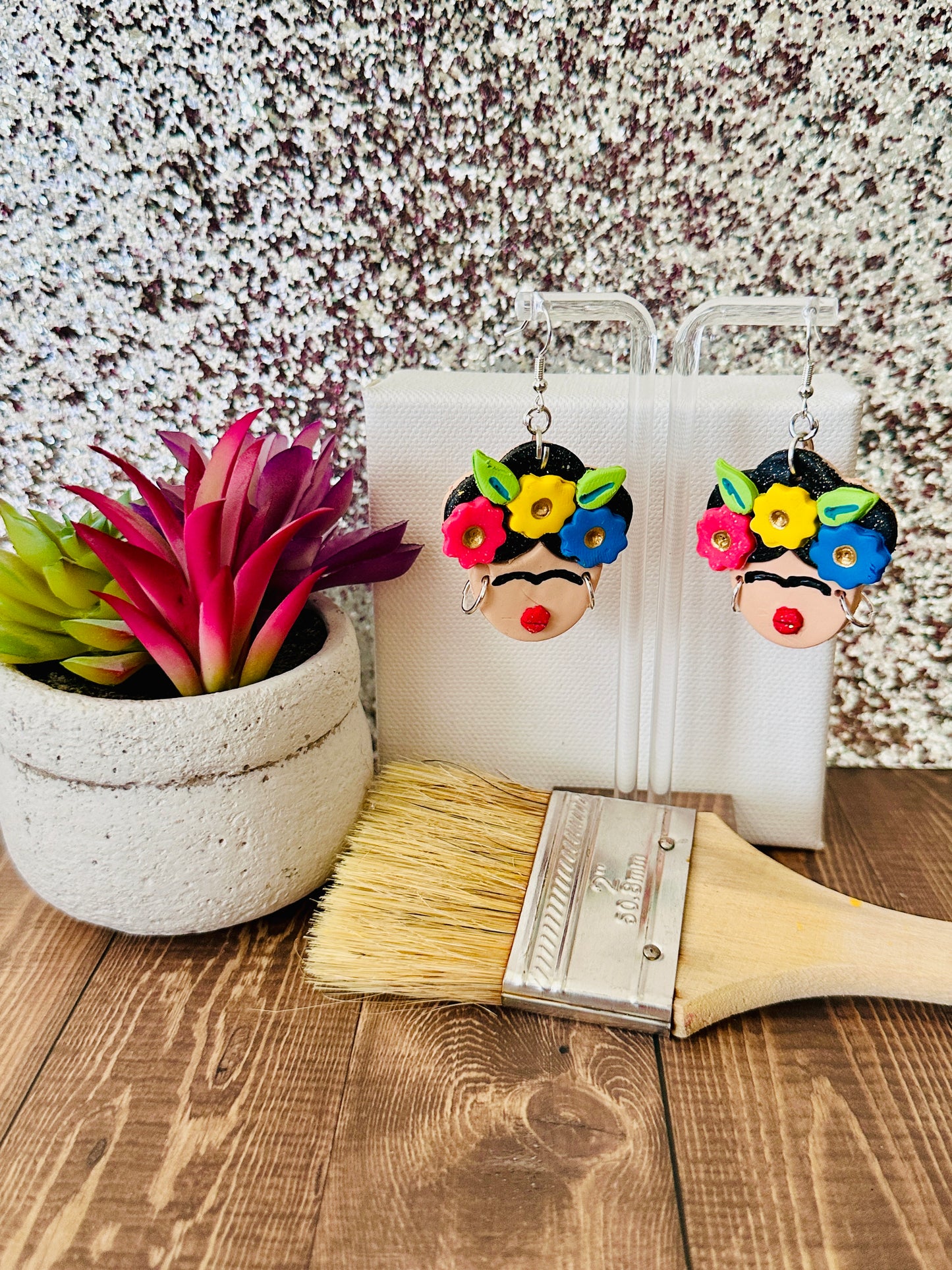 Large Frida Kahlo Bold Flower Crown polymer clay earrings with colorful floral crown hanging on a clear earring stand next to a potted plant and paintbrush on a wooden surface.