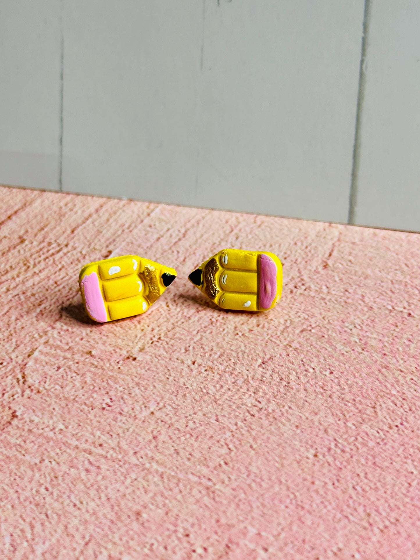 Am angled view of a set of pencil stud earrings on a pink table The stud is yellow with a pink eraser, white shadows, gold colored wood, and black pencil tip.