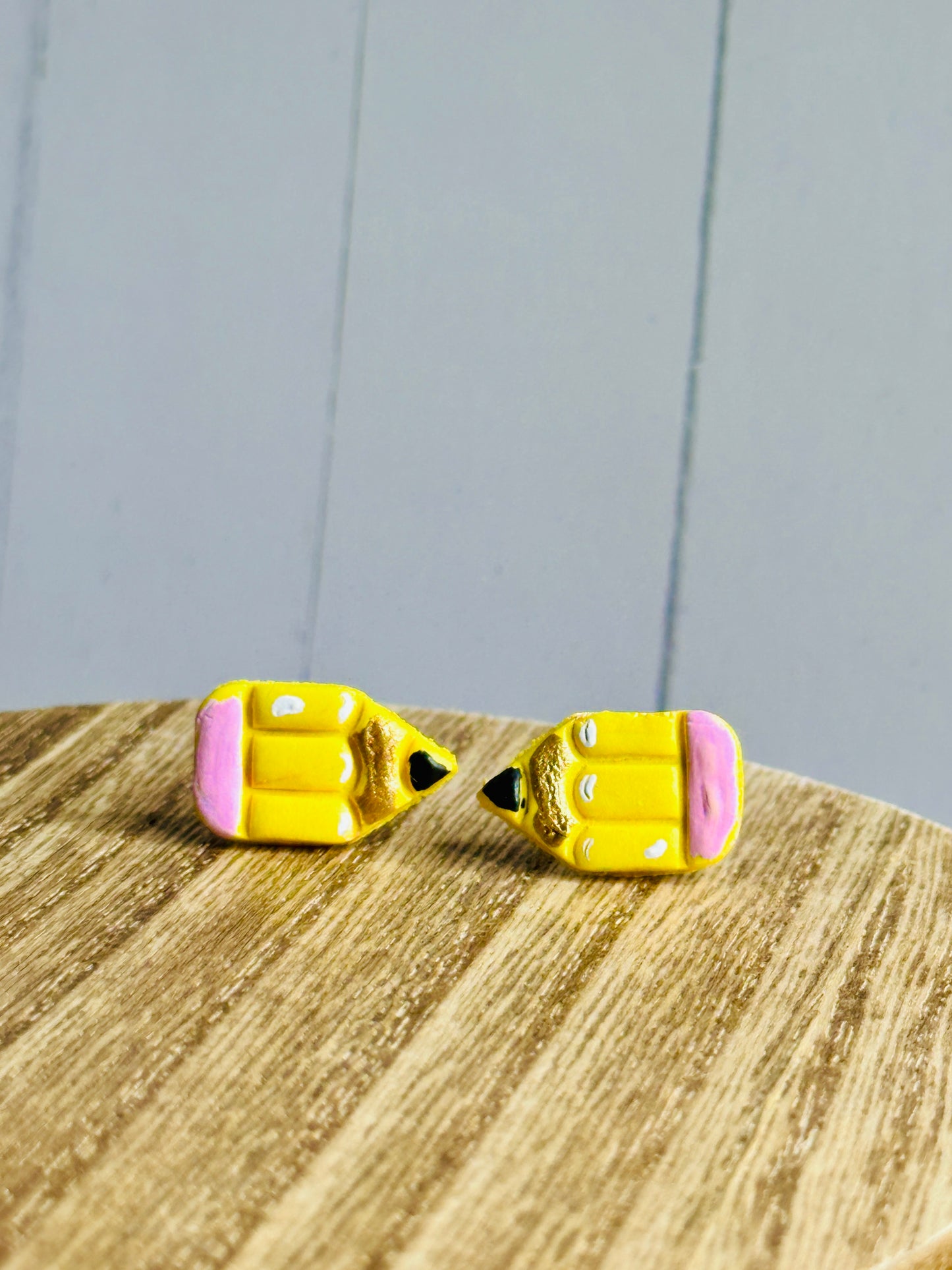 A set of pencil stud earrings on a wooden table The stud is yellow with a pink eraser, white shadows, gold colored wood, and black pencil tip.
