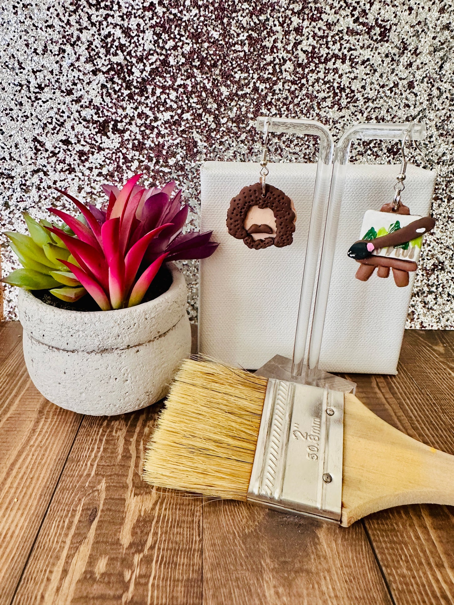 Small Bob Ross earring set displayed by a paint canvas with a paintbrush and a plant on a wooden surface.