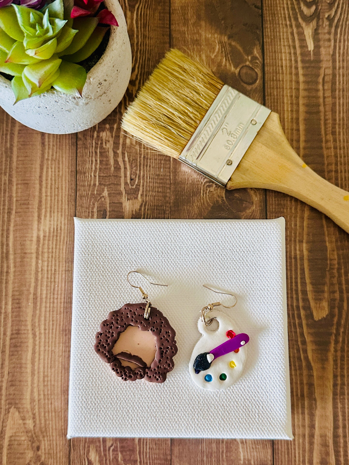 Large Bob Ross earring set displayed by a paint canvas with a paintbrush and a plant on a wooden surface.