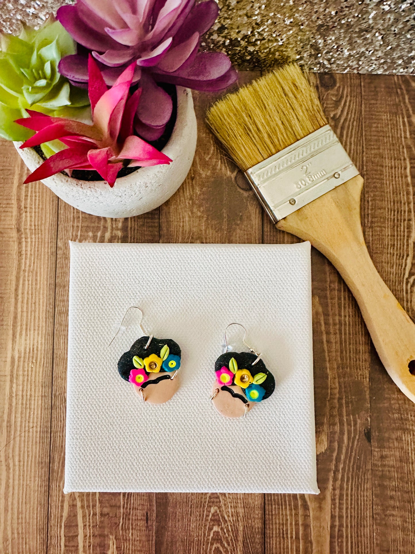 The small variant of Frida Kahlo-themed earrings on a white paint canvas with a paintbrush and flowers in the background and sitting on a wooden table.