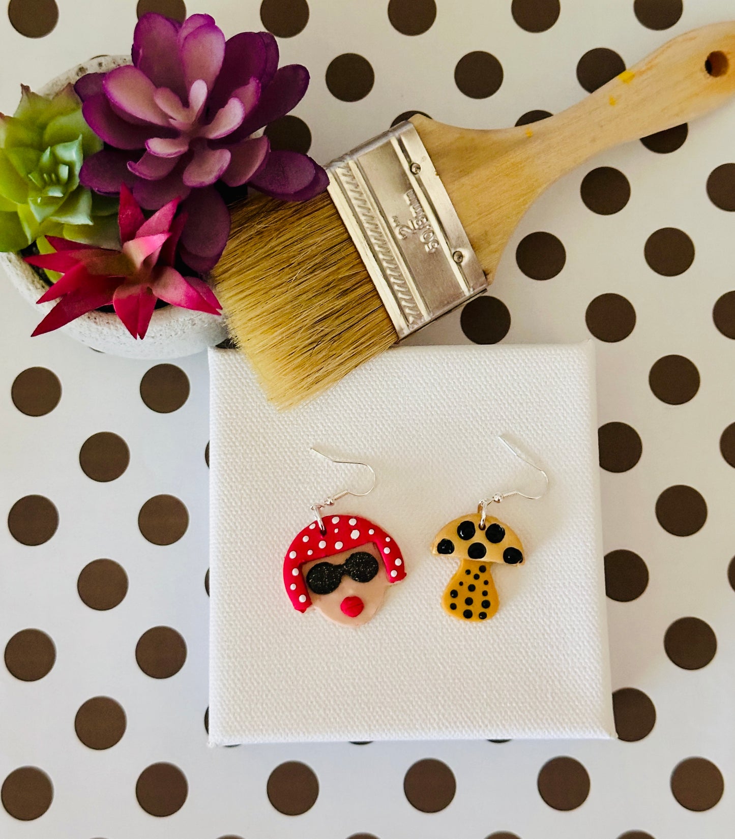 The small variant of the Yayoi Kusama polymer clay earrings displayed on white canvas with paintbrush and potted plant on black and white polka dotted table, perfect for art teachers.