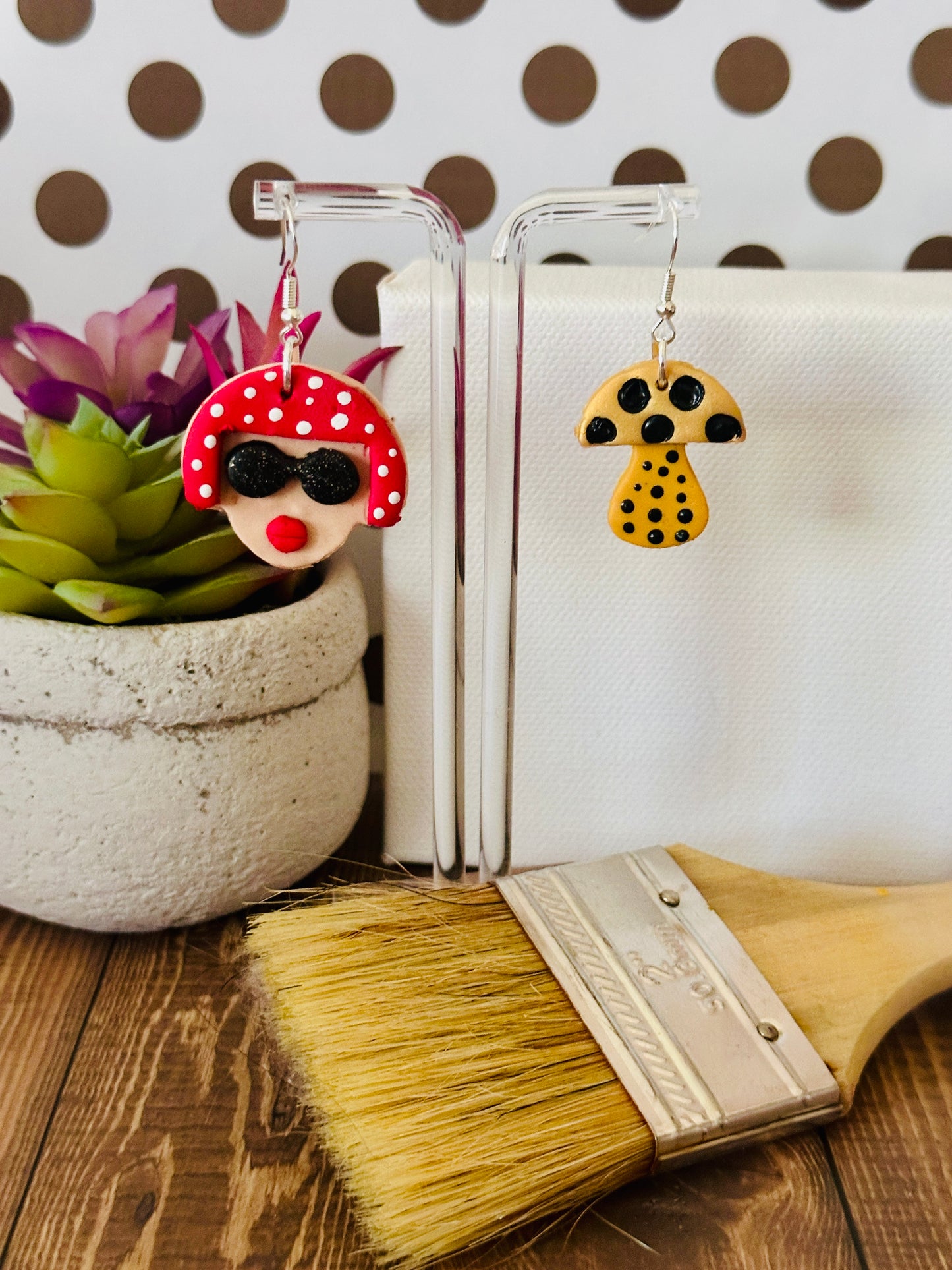 Small variant of the Yayoi Kusama inspired earrings hanging on clear earring stand with polka dot background and paintbrush, showing artistic portrait and mushroom design.