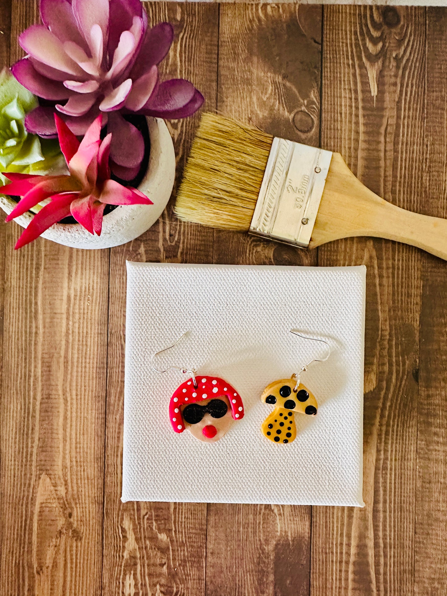 Small Yayoi Kusama polymer clay earrings with portrait featuring red polka dot hair, black sunglasses and yellow mushroom on blank canvas and wooden table.