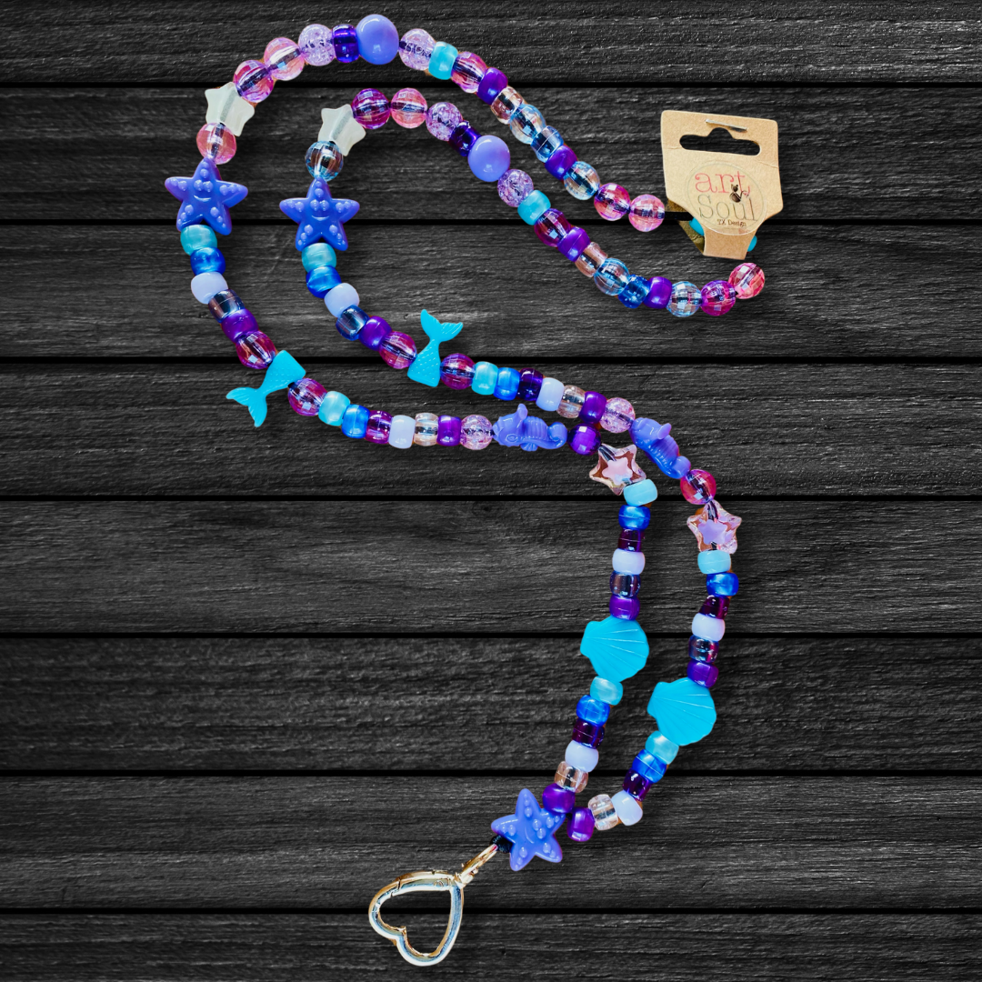 The Under The Sea Mermaid Lanyard on a dark black wooden background that contrasts the deep sea blue color of the beads. Starfish, mermaid tails, seashells, and sea horse beads decorate this lanyard.