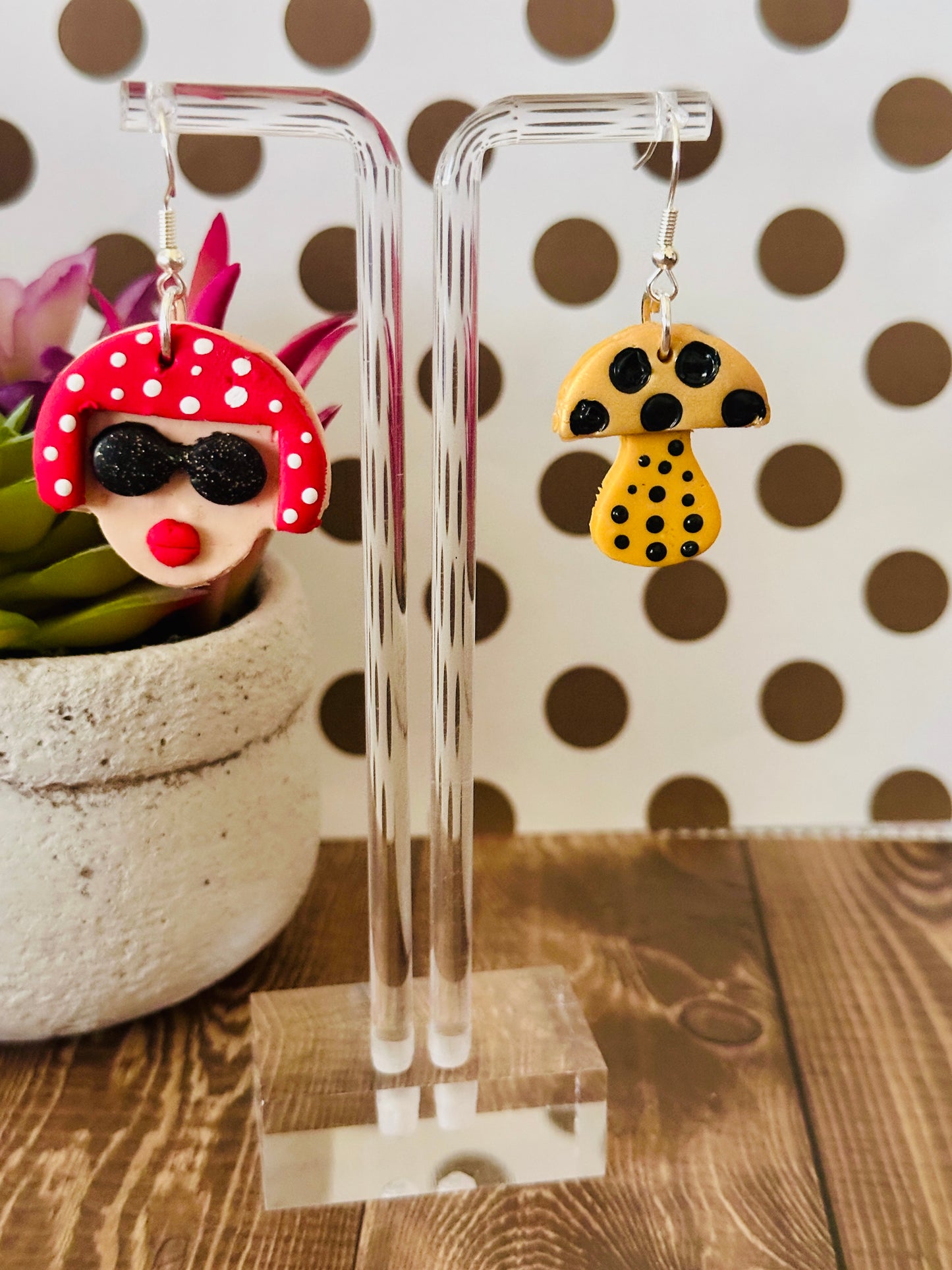 Close-up of Yayoi Kusama polymer clay earrings on clear stand with polka dot background and wooden table, highlighting handcrafted details.