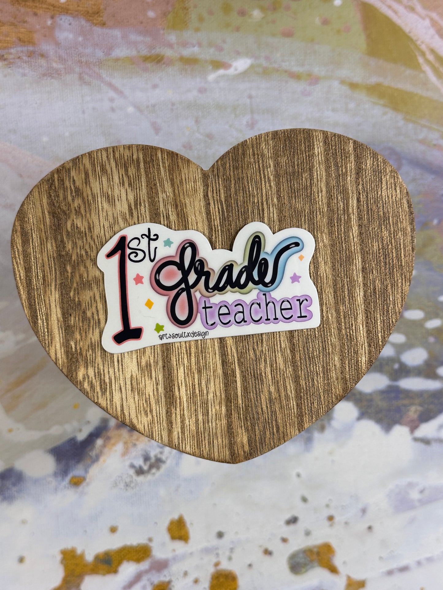 1st Grade teacher vinyl sticker on a wooden heart-shaped table with colorful background, showing bold cute font with colorful letter highlights.