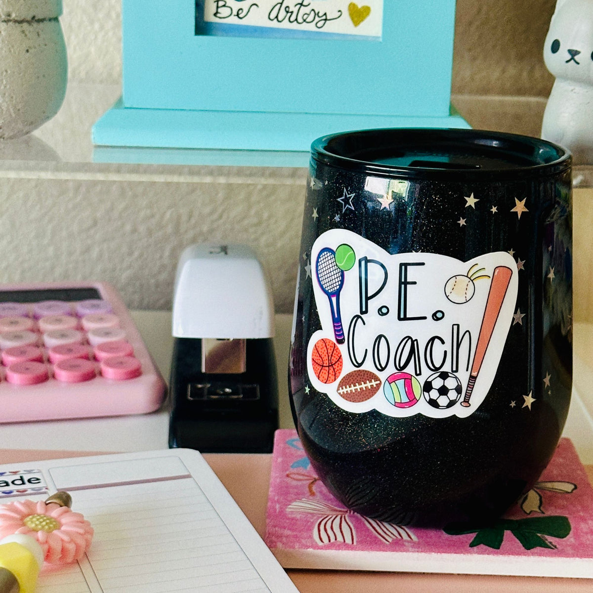A close-up of a black glitter tumbler cup with a “P.E. Coach” Vinyl Sticker