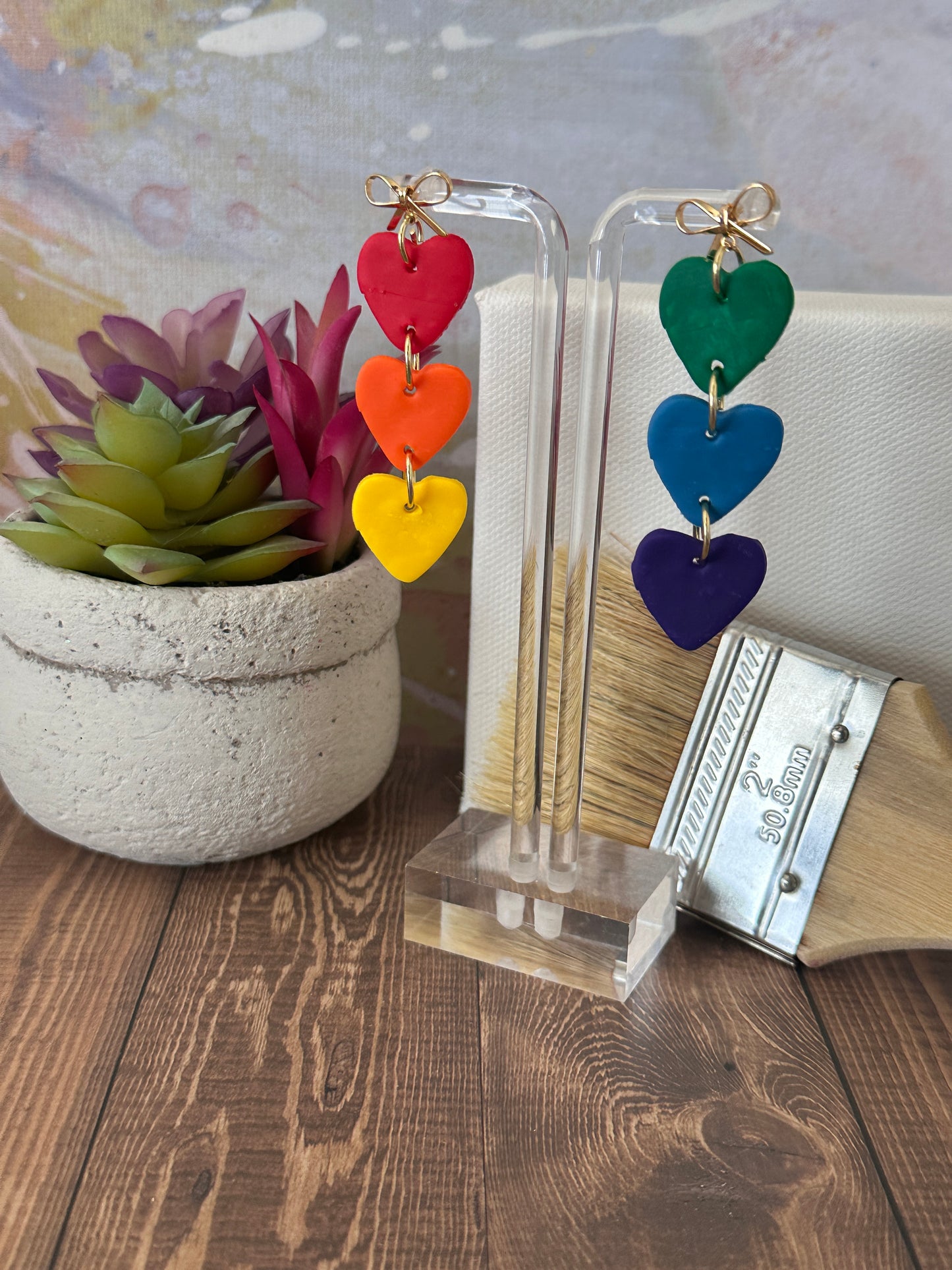 Colorful heart-shaped earrings on a clear stand with a plant and book in the background.
