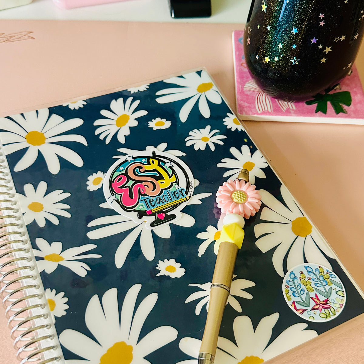 A spiral-bound notebook with a navy blue cover decorated with a white daisy floral pattern, featuring a colorful 'ESL Teacher' sticker on the front. A pink flower-shaped pen with a gold handle rests on top of the notebook, set against a peach-colored desk background with other desk accessories.