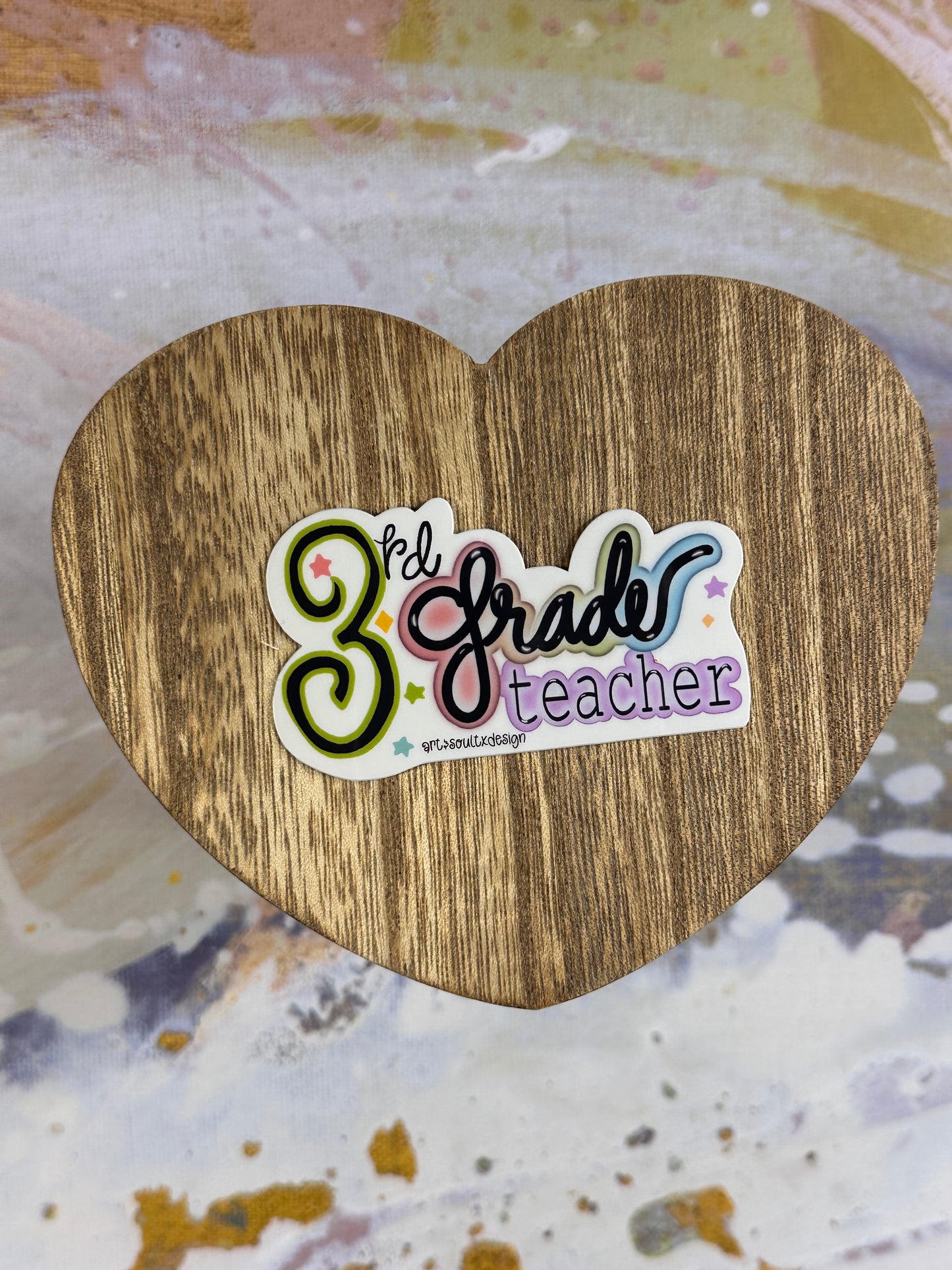 3rd Grade teacher vinyl sticker on a wooden heart-shaped table with colorful background, showing bold cute font with colorful letter highlights.