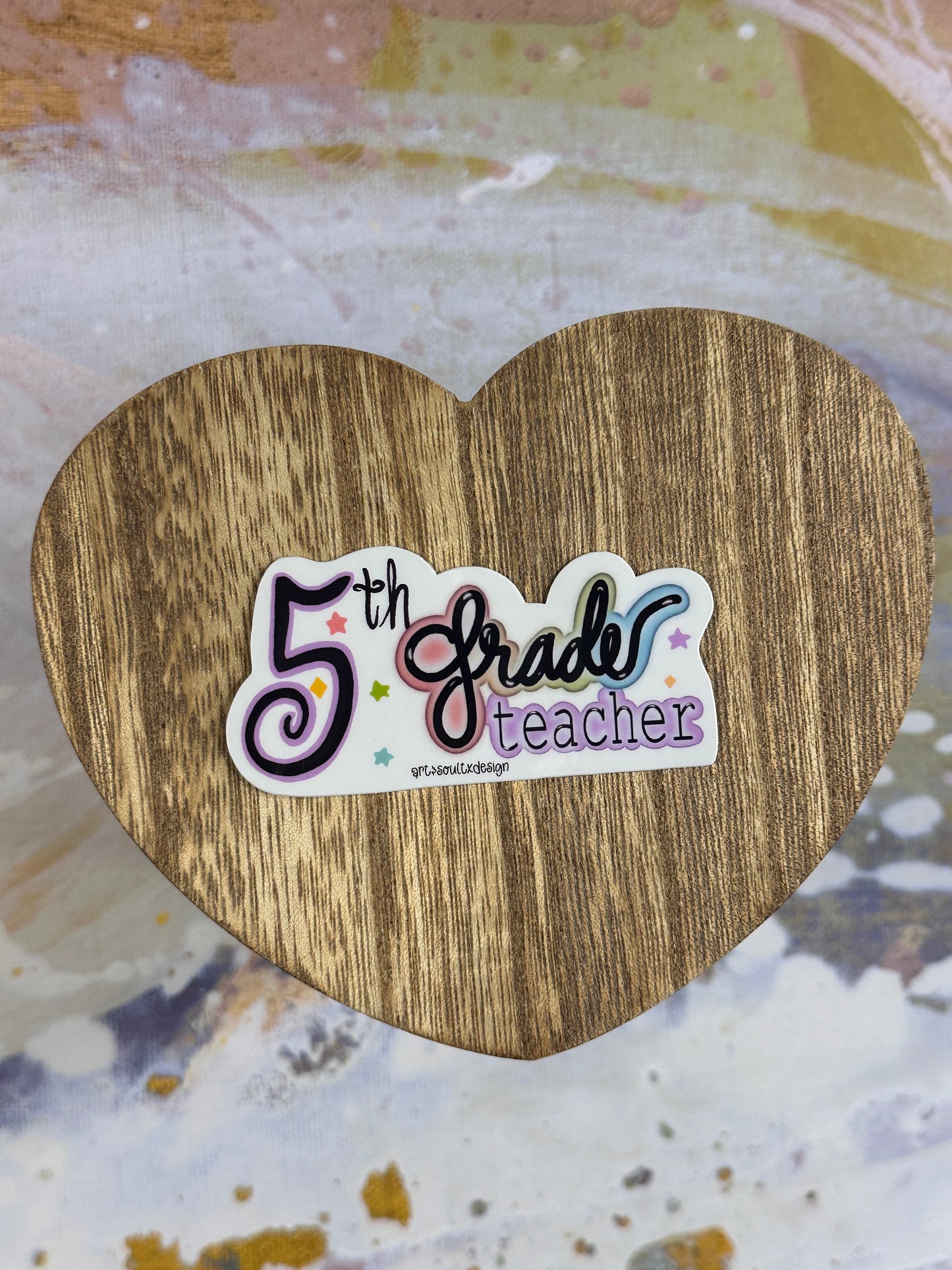 5th Grade teacher vinyl sticker on a wooden heart-shaped table with colorful background, showing bold cute font with colorful letter highlights.