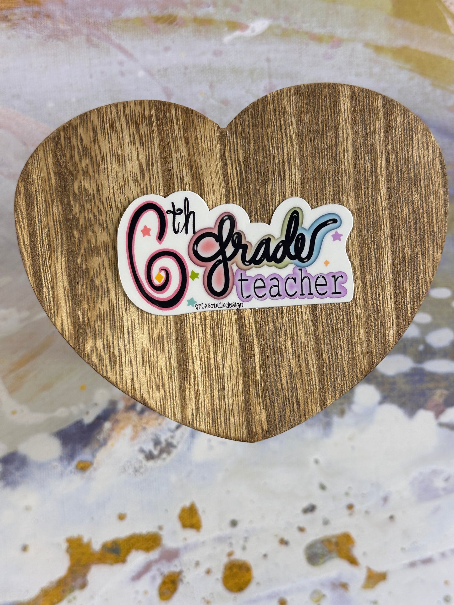 6th Grade teacher vinyl sticker on a wooden heart-shaped table with colorful background, showing bold cute font with colorful letter highlights.