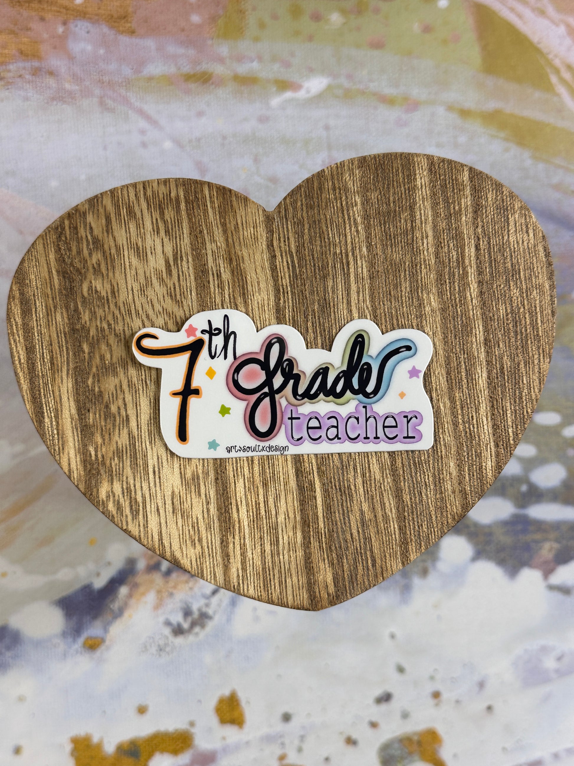 7th Grade teacher vinyl sticker on a wooden heart-shaped table with colorful background, showing bold cute font with colorful letter highlights.