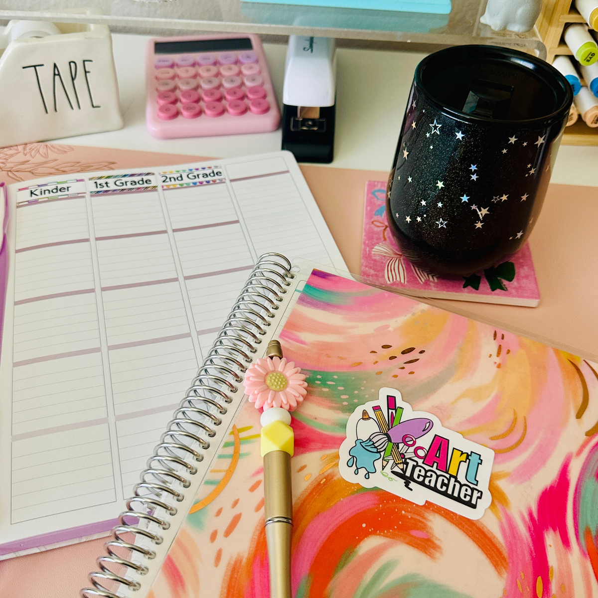 A styled desk setup featuring a weekly planner page, a spiral notebook with a colorful abstract cover and a 'Art Teacher' sticker, a black starry tumbler, and other stationery items.