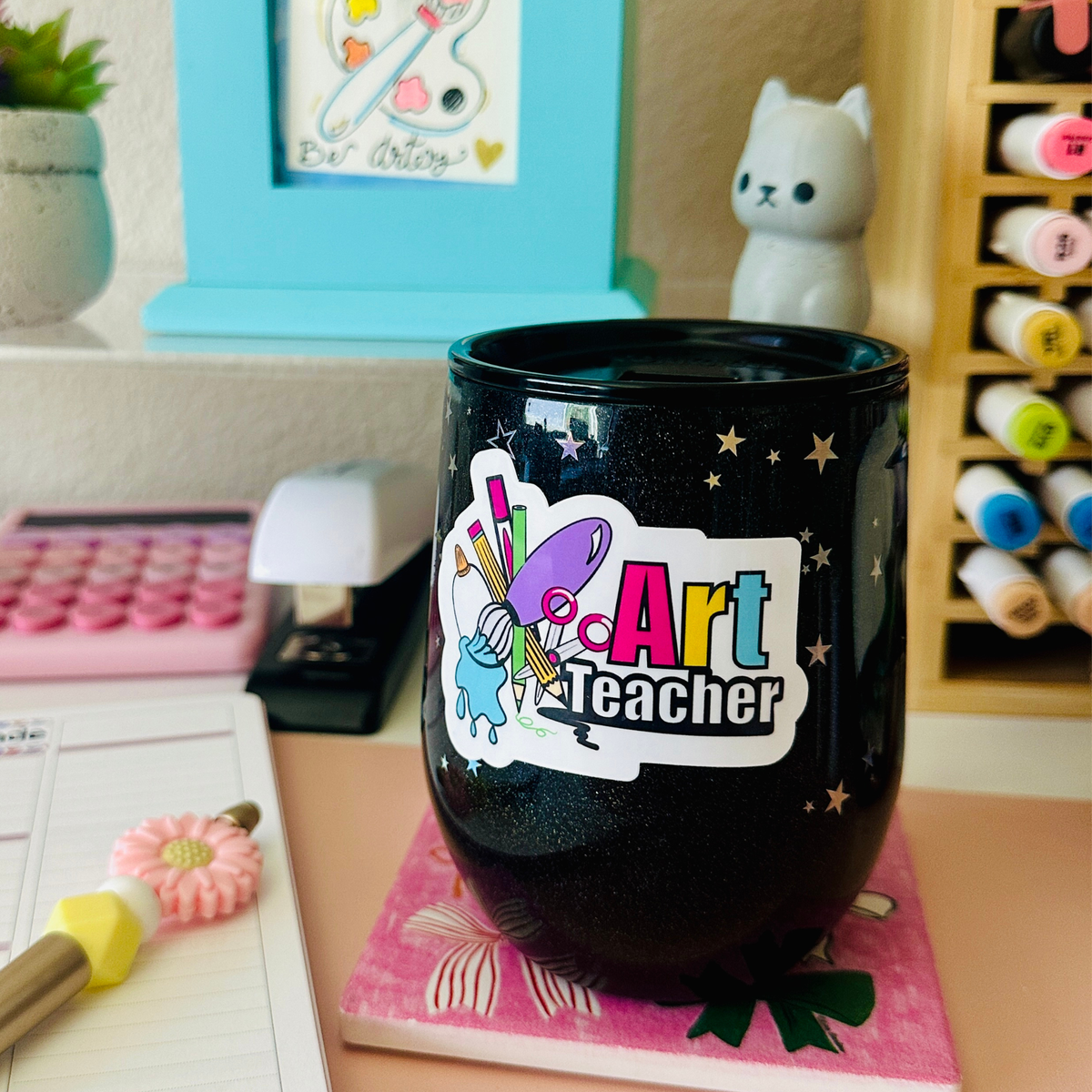 A black glitter stemless wine tumbler with a 'Art Teacher' sticker, set on a pink bow coaster in a desk environment.