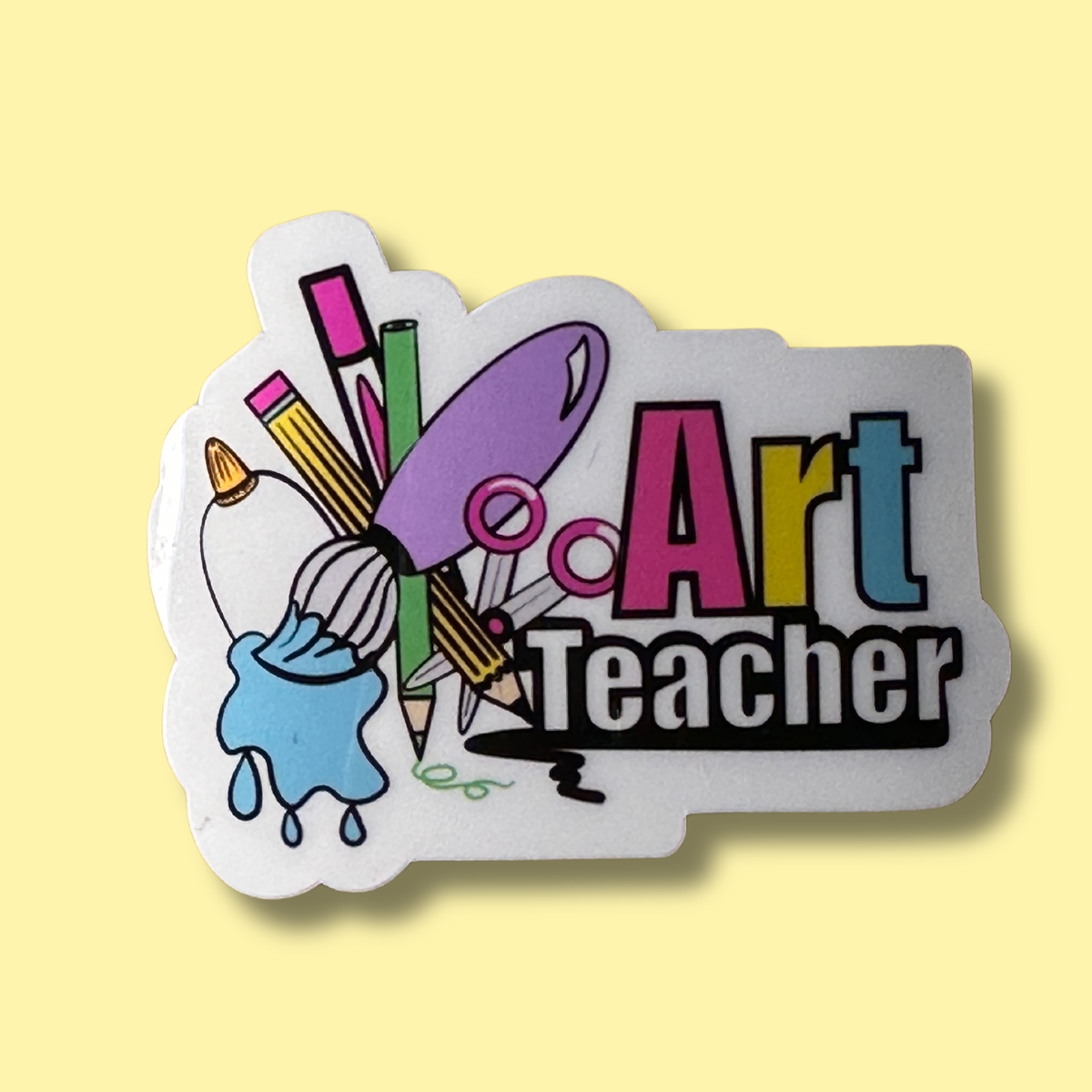 A die-cut sticker featuring the words 'Art Teacher' in colorful block letters with illustrated art supplies including a paintbrush, paint bucket, paint palette, pencils, and scissors on a white background, set against a pale yellow backdrop.