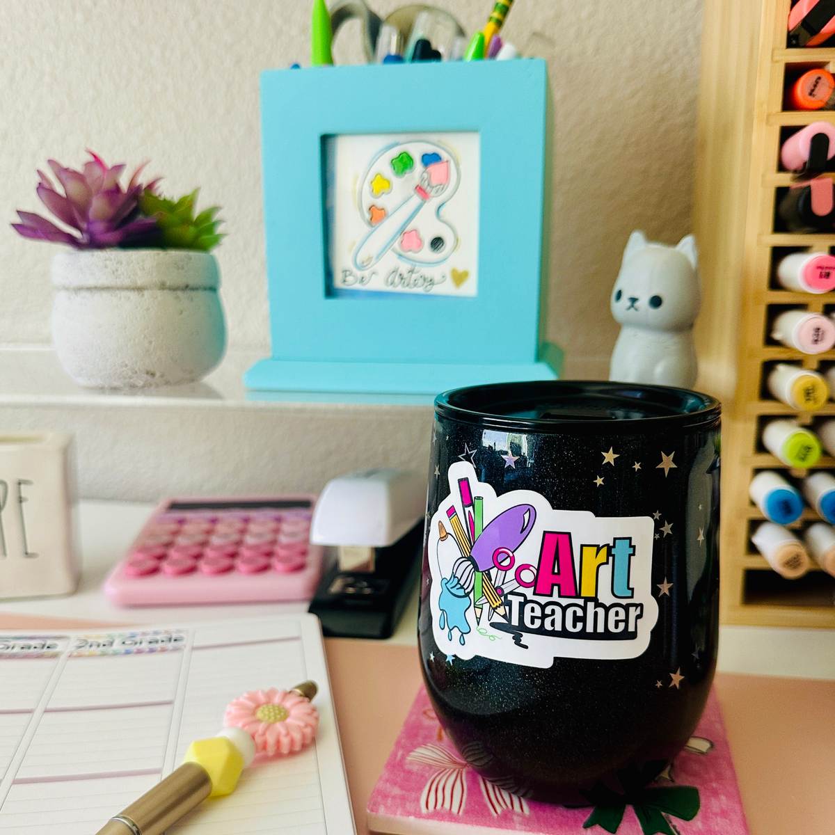A black glittery stemless tumbler cup with a colorful 'Art Teacher' sticker, placed on a desk surrounded by art supplies, a small plant, and stationery.