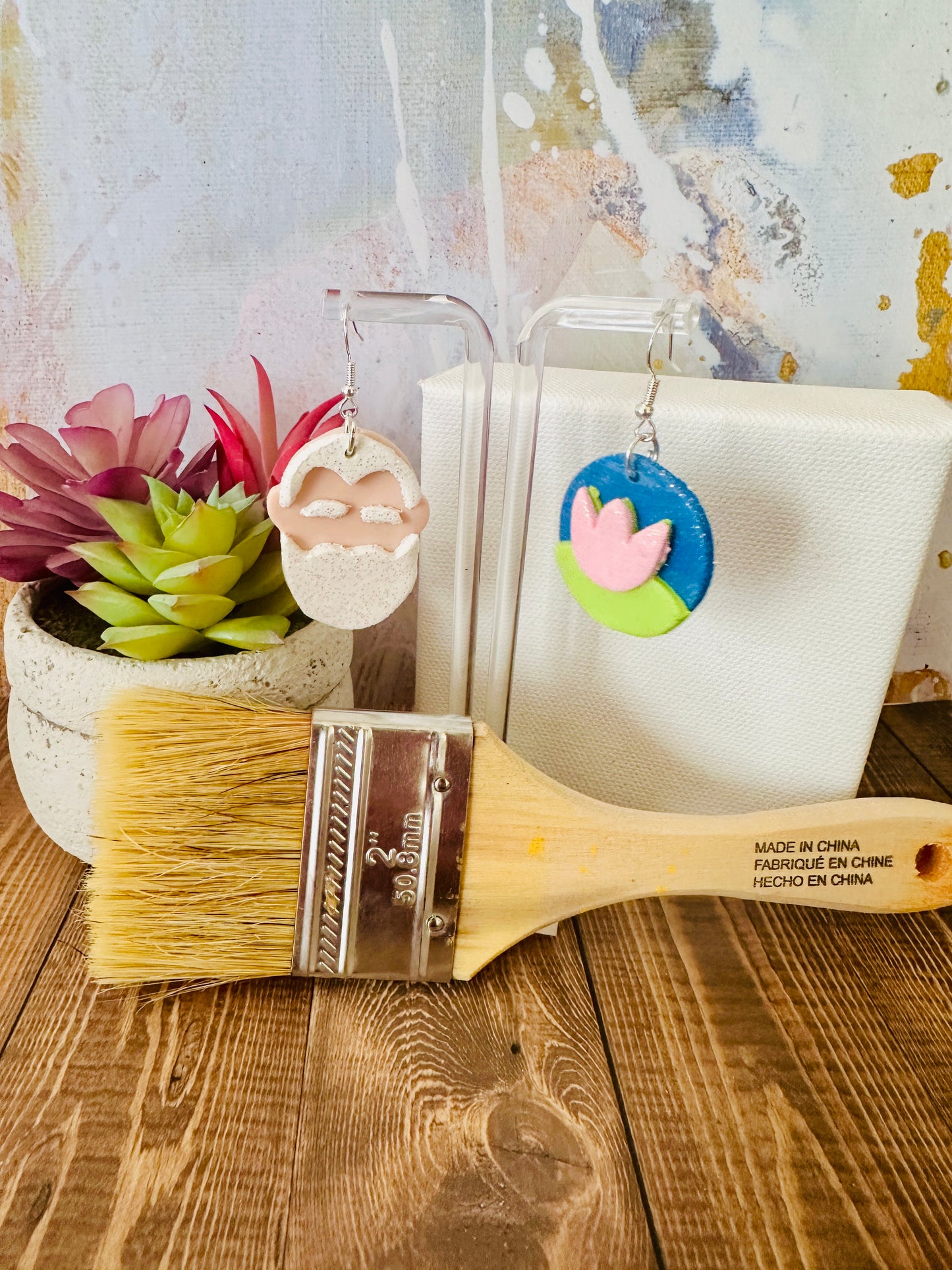 Handcrafted Monet earrings on stand with small white canvas, paintbrush, plants on wood table, colorful backdrop.