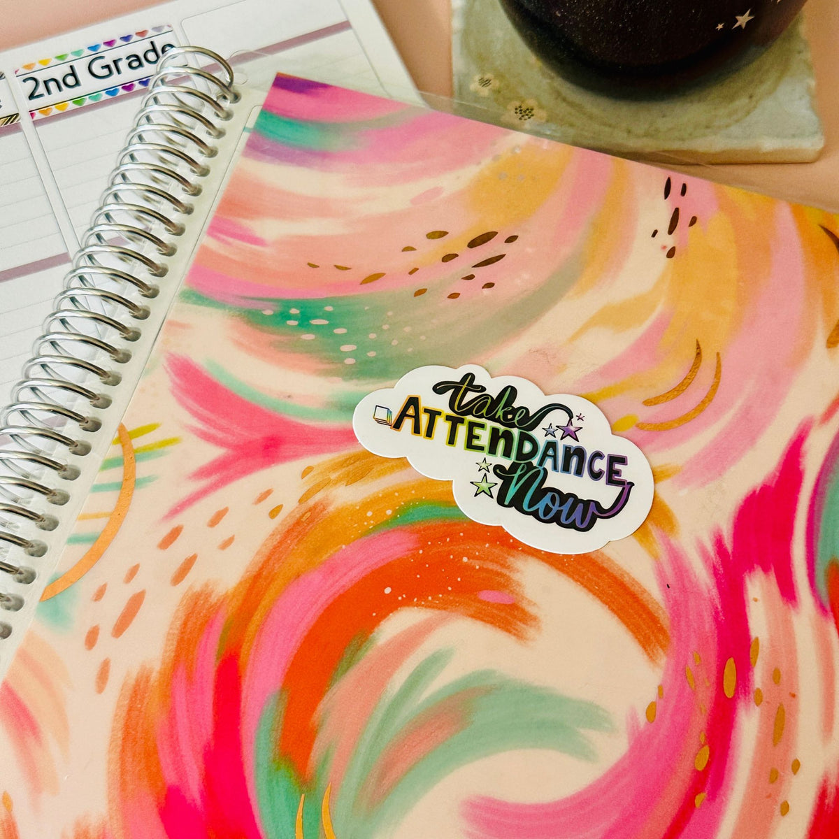 A spiral-bound notebook with a colorful abstract watercolor cover featuring pink, orange, yellow, and teal swirls. A die-cut sticker on the cover reads 'take ATTENDANCE NOW' in stylized lettering.