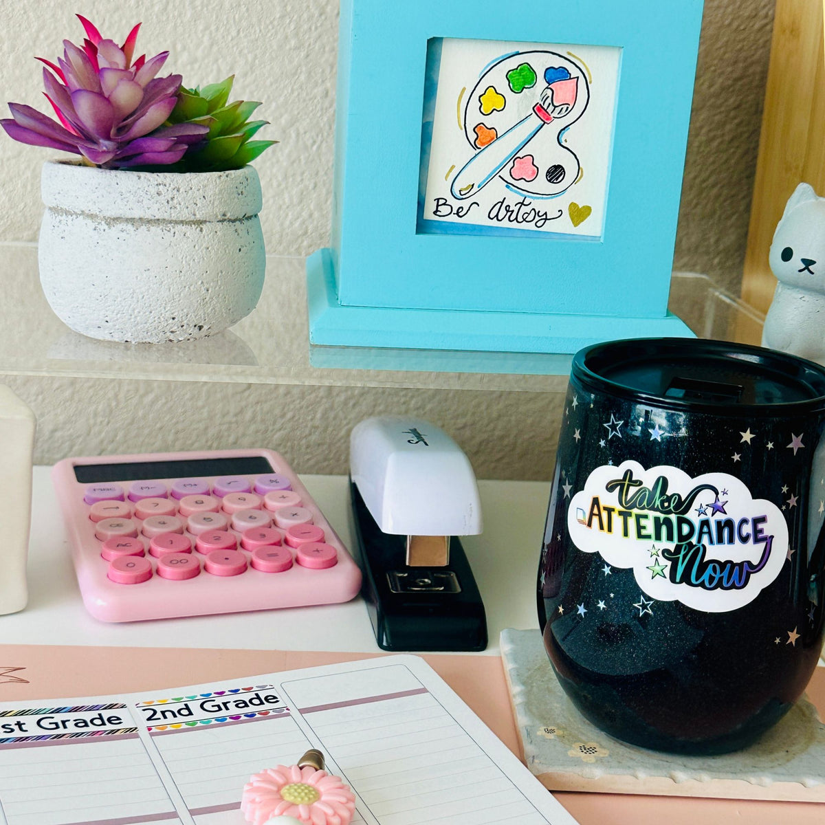 A styled desk scene featuring a black glittery tumbler cup with 'take ATTENDANCE Now' printed on it, alongside a pink calculator, white stapler, planner page, small succulent in a textured pot, and a turquoise framed art print.