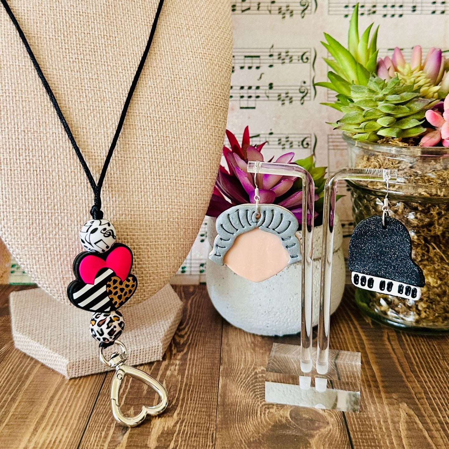 Necklace with decorative pendant on a wooden surface with plants and music sheet background along with a pair of earrings on a clear acrylic stand on the right side.