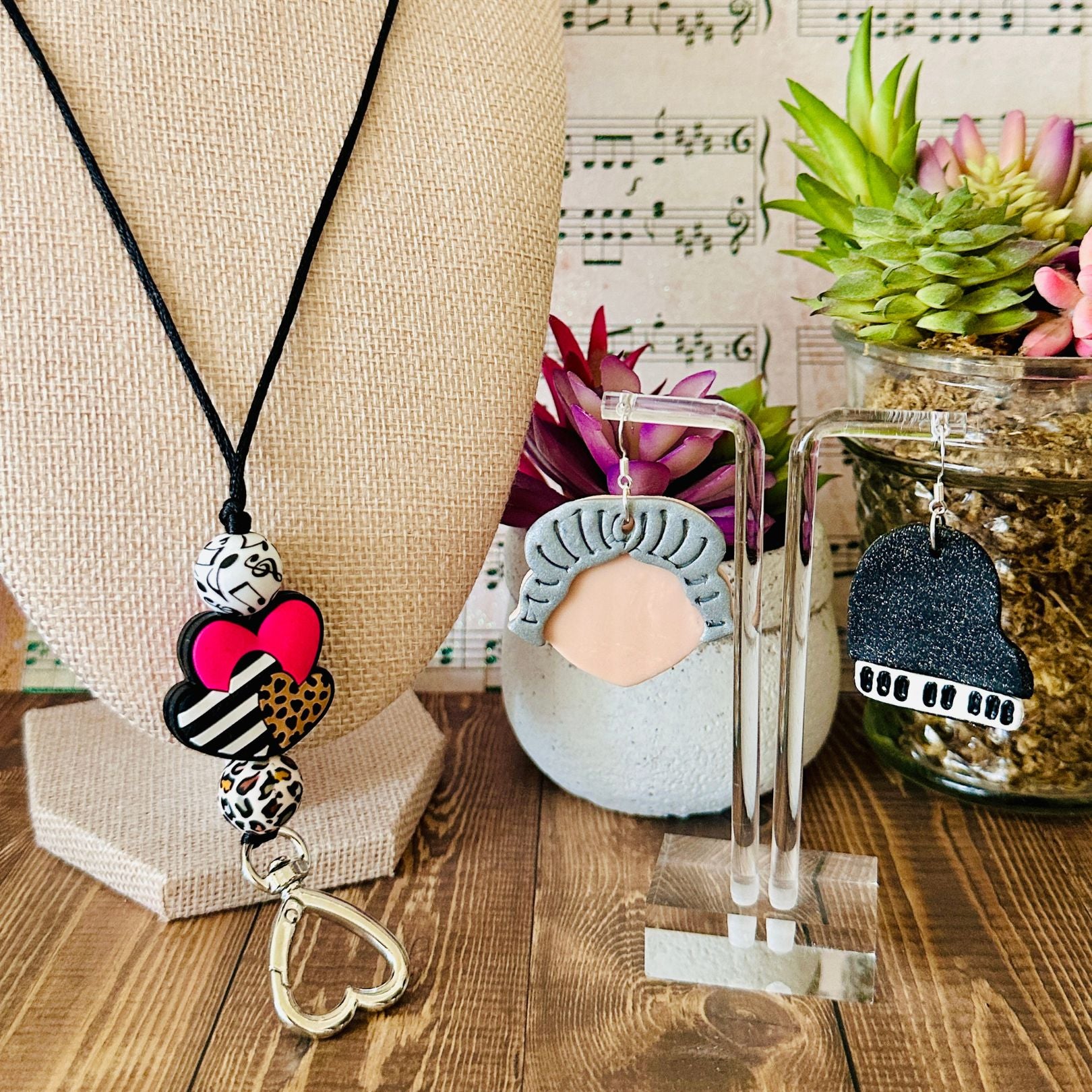 Necklace with decorative pendant on a wooden surface with plants and music sheet background along with a pair of earrings on a clear acrylic stand on the right side.