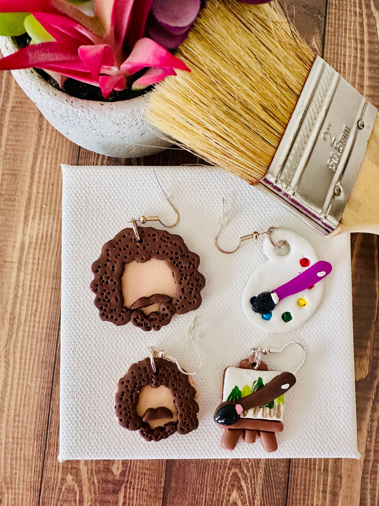 A top view of Bob Ross polymer clay earrings shown so shoppers can compare details between large and small sizes.  The large variant has a paint palette and the small variant has a canvas painted with cute little trees.