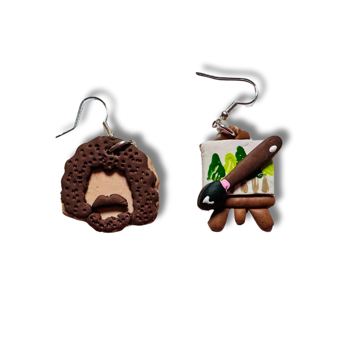 Handmade Bob Ross Style Polymer Clay Earrings.  The earring on the left is a 1x 1 inch Bob Ross Head shaped with beige skin, brown hair, beard, and mustache. The earring on the right is a 0.75 x 1 inch paint canvas with a paintbrush on top. The canvas shows a brown easel holding a white canvas with hand painted green trees. On top of the canvas is a brown paint brush with black brush and shine of white to reflect the lighting.  These earrings have nickel free silver hooks. 