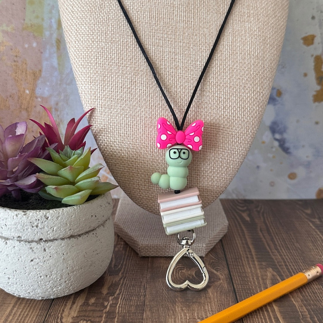 Close up of lanyard with a green book worm, pink and white bow, and books pendant on a mannequin bust with a plant and pencil in the foreground.