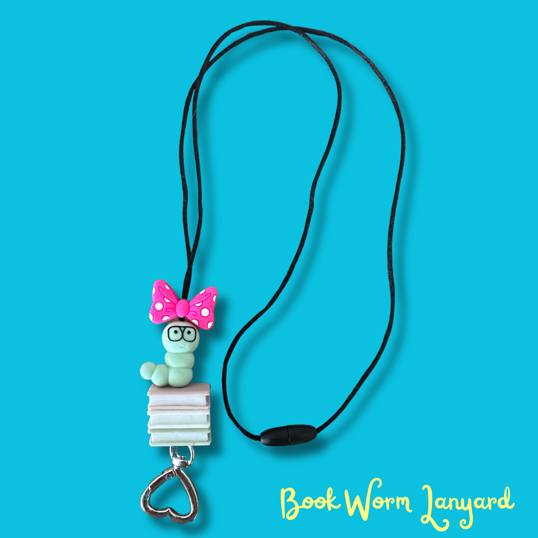 A black cord necklace featuring a charm shaped like a light green bookworm wearing a pink polka-dot bow and glasses, sitting on a stack of books and hanging from a silver heart-shaped clasp, set against a bright blue background with 'Book Worm Lanyard' text in the lower right.