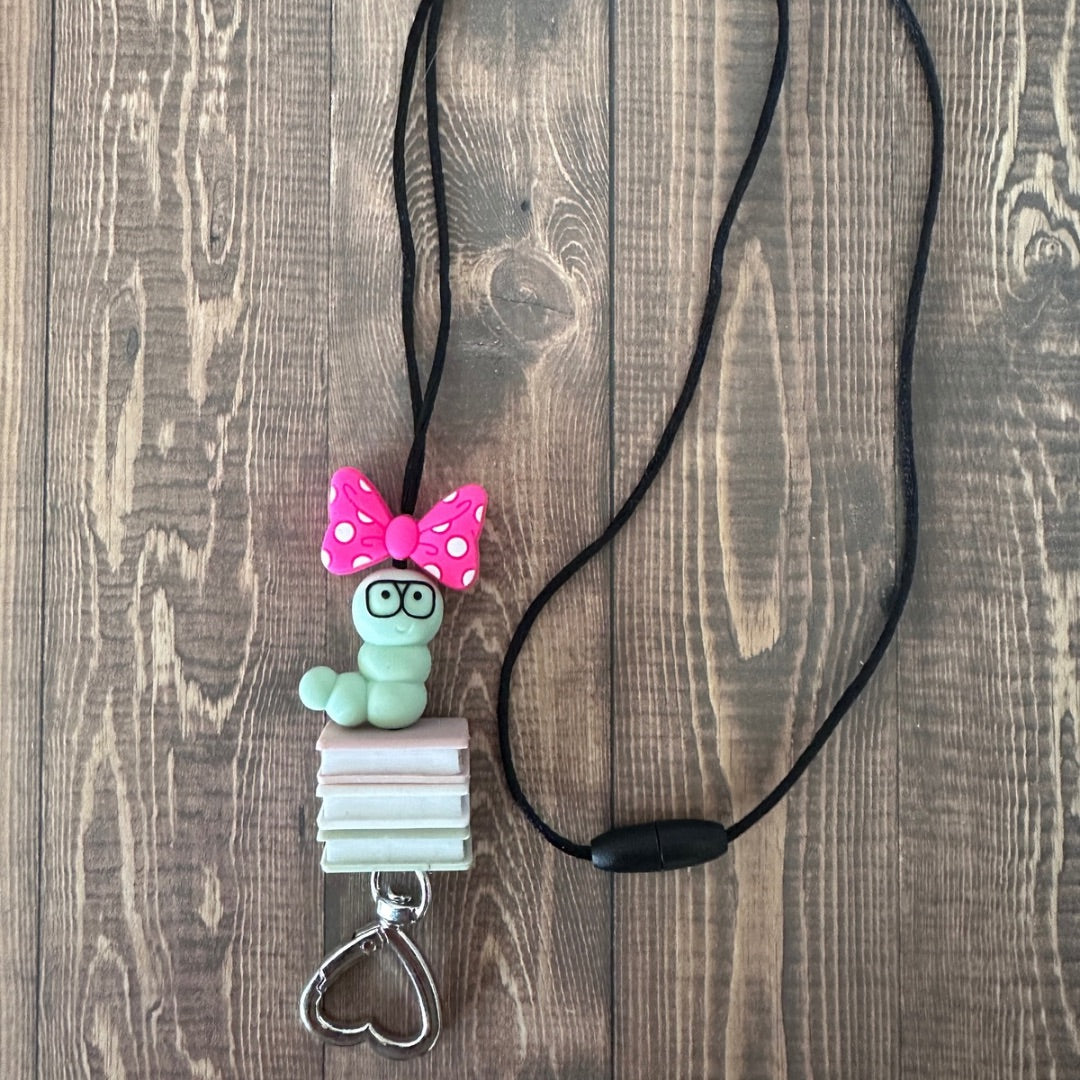 Lanyard on wooded background showing the close up of silicone beaded pink bow, green worm, pink book, white book, and green book.  