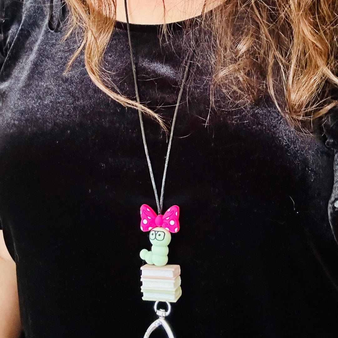 Lanyard with a green book worm, pink bow, and 3 book silicone beads on a person modeling  with black shirt. 