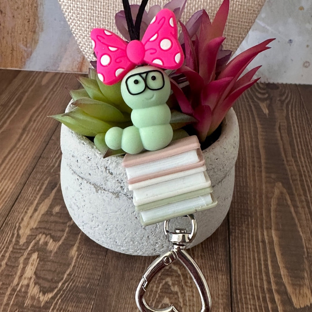 Super close up of Book worm lanyard resting on a potted succulent plant.  It shows the details of each silicone beads from a pink and white polka dotted bow, green worm with glasses, pink book, white book, green book, and a heart clasp to hold keys and badge card.